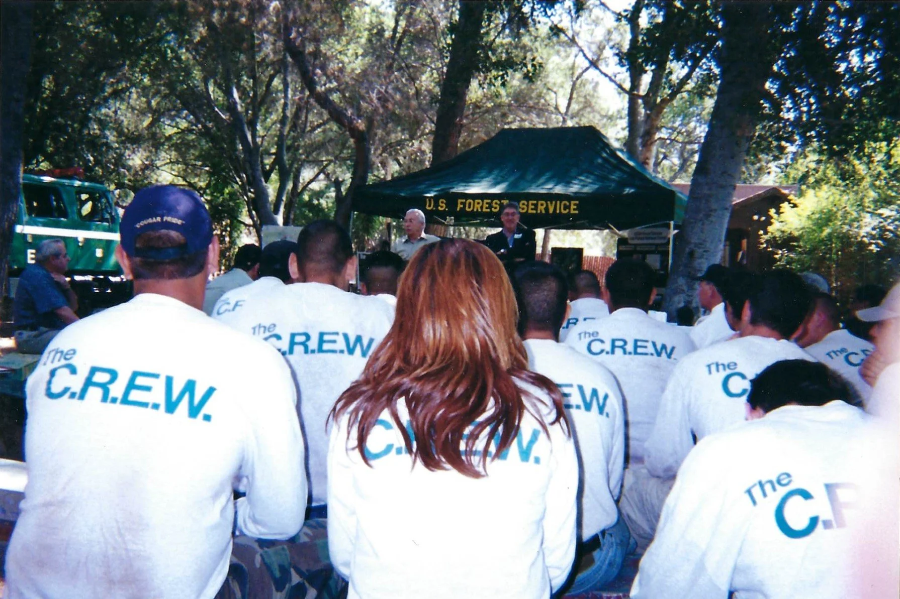 People wearing white shirts with 'The C.R.E.W.' printed on the back gathered outdoors for a presentation or meeting under a black canopy with yellow text that reads 'U.S. Forest Service' amidst trees.