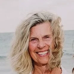Close-up of a woman with gray, curly hair smiling outdoors near the water