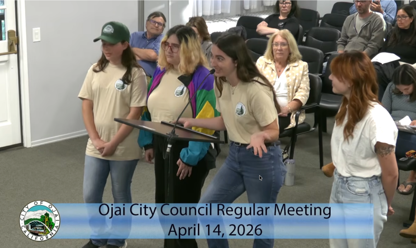 A woman speaking at a podium during the Ojai City Council regular meeting on April 14, 2026, with five young women standing next to her. Several people are seated in the audience behind them. The event appears to be in a council chamber.