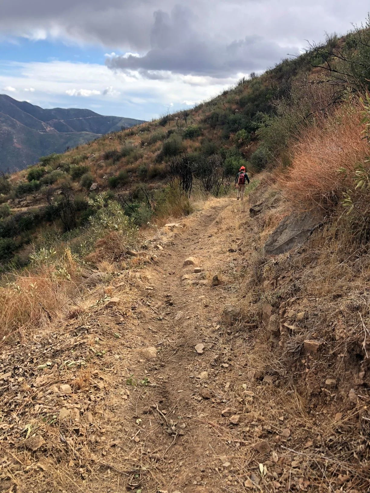 A dirt trail winding through a dry landscape with sparse shrubs and bushes, surrounded by mountainous terrain under a cloudy sky. A person wearing a red hat and backpack is seen walking along the trail.