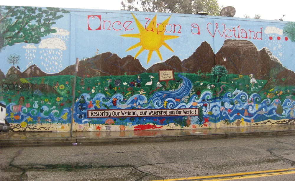 A wall mural titled Once Upon a Wetland that paints the Ojai Meadows Preserve, with mountains, a sun, a creek, an egret and children planting, over the words Restoring Our Wetland, our Watershed and Our World!