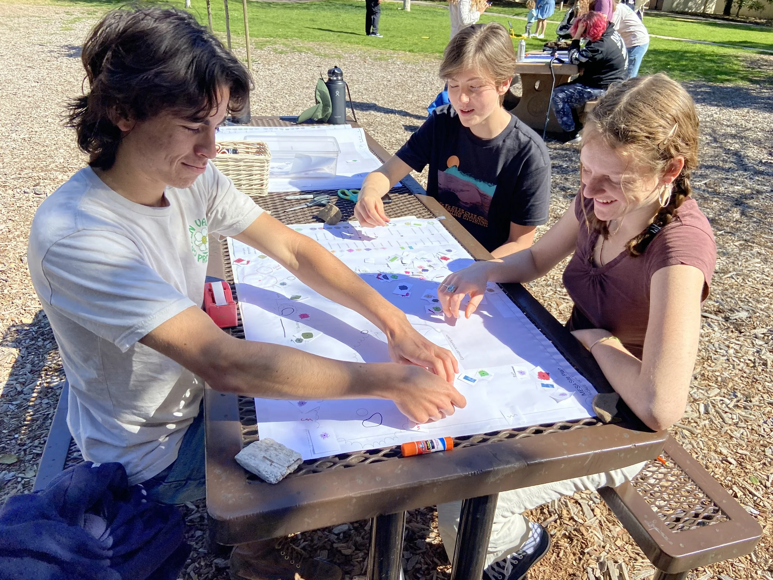 Three people sitting at an outdoor table having a conversation, with hands resting on a sheet of paper, in a park-like setting on a sunny day.