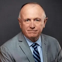 A middle-aged man with a serious expression, wearing a light gray suit, blue shirt, and striped tie, against a dark background.