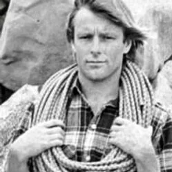Black and white photo of a young man with medium-length hair holding a striped scarf around his neck, standing outdoors with rocks in the background.
