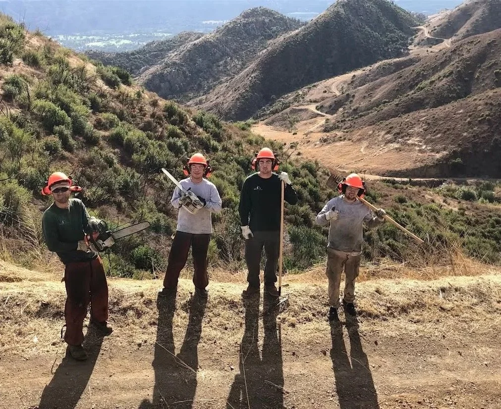 Four workers wearing orange helmets, gloves, and work clothes standing on a dirt trail in a mountainous area, some holding tools like chainsaws, shovels, and pickaxes.