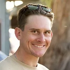 A young man smiling outdoors with sunglasses on his head and a blurred background of trees.