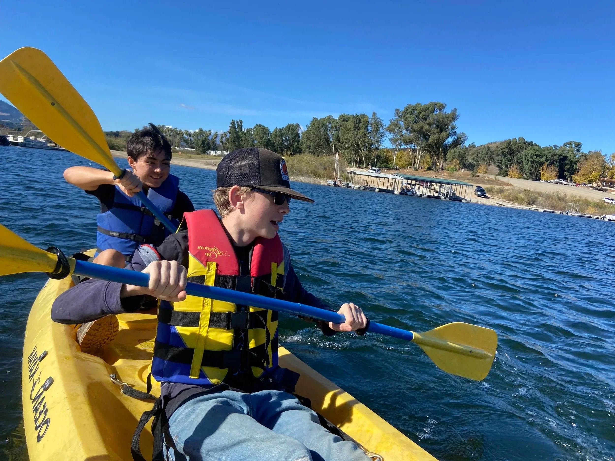 Two boys kayaking on a lake with trees and a shoreline in the background under a clear blue sky.