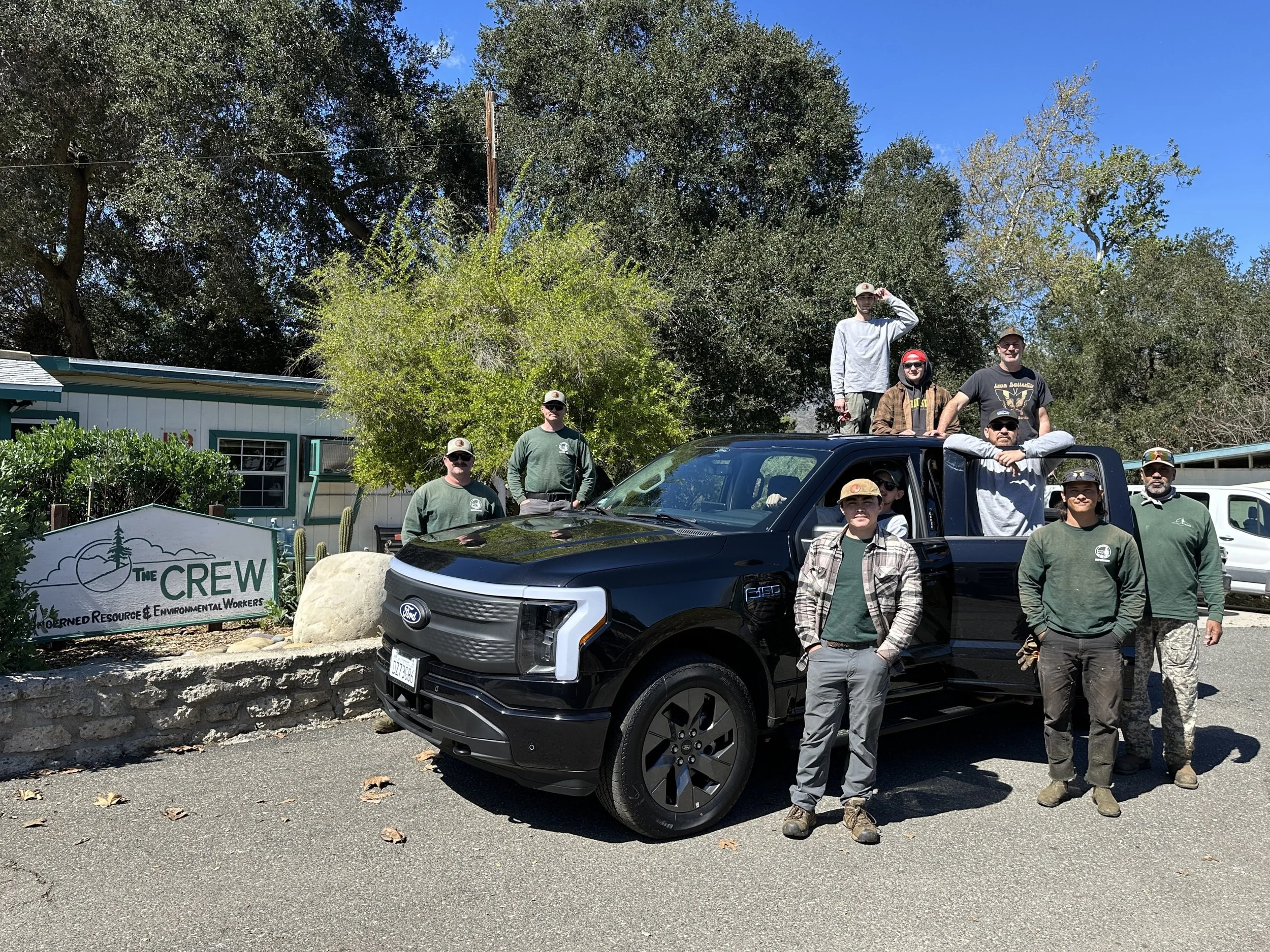 A group of people posing outdoors in front of trees and a building, with several standing and some sitting on a black pickup truck, in a sunny environment.