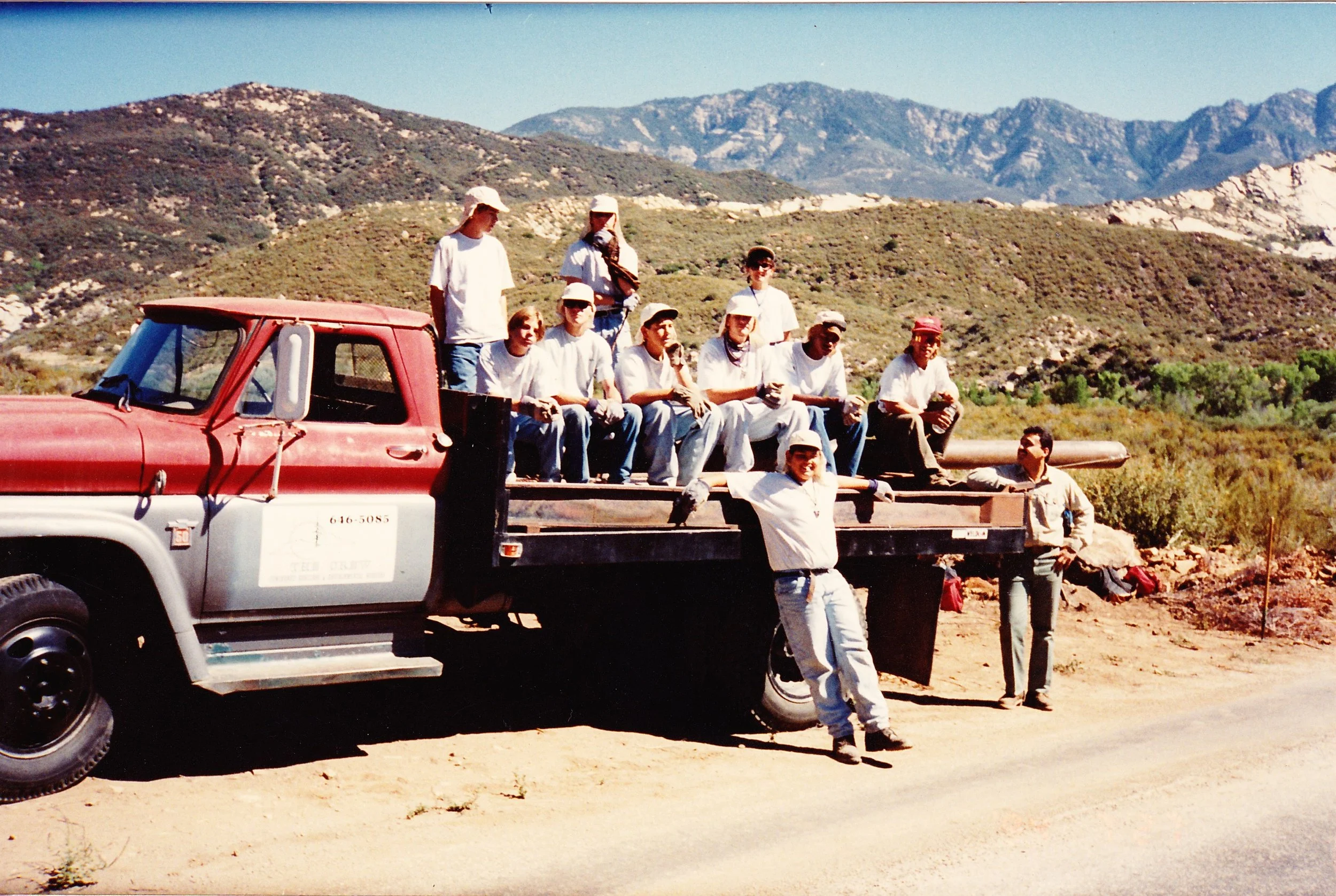 A group of people sitting on the back of a red and white pickup truck in a mountainous landscape with clear blue skies.