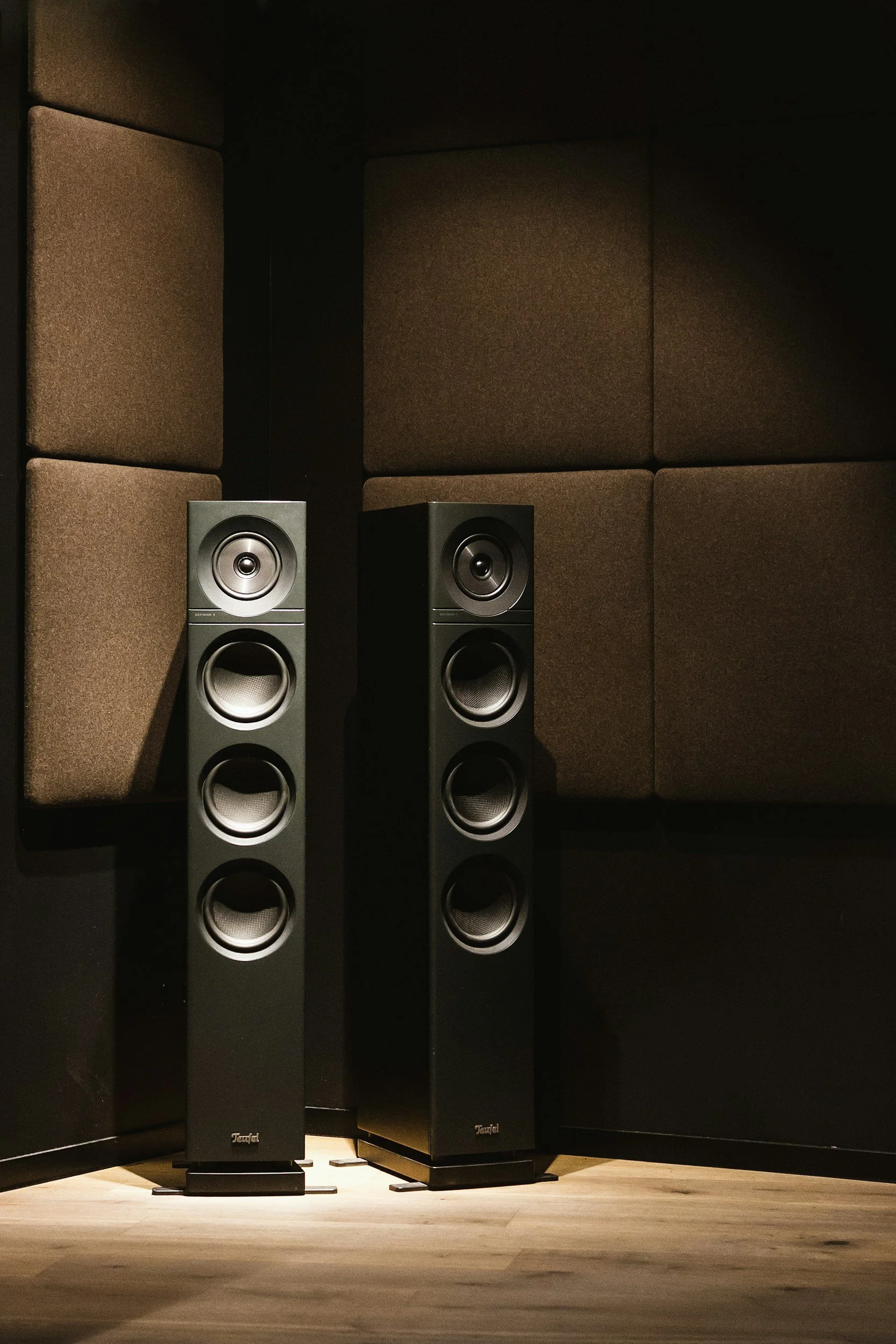 Two tall black speakers with multiple drivers are in front of soundproof brown panels arranged as walls in a dark room.