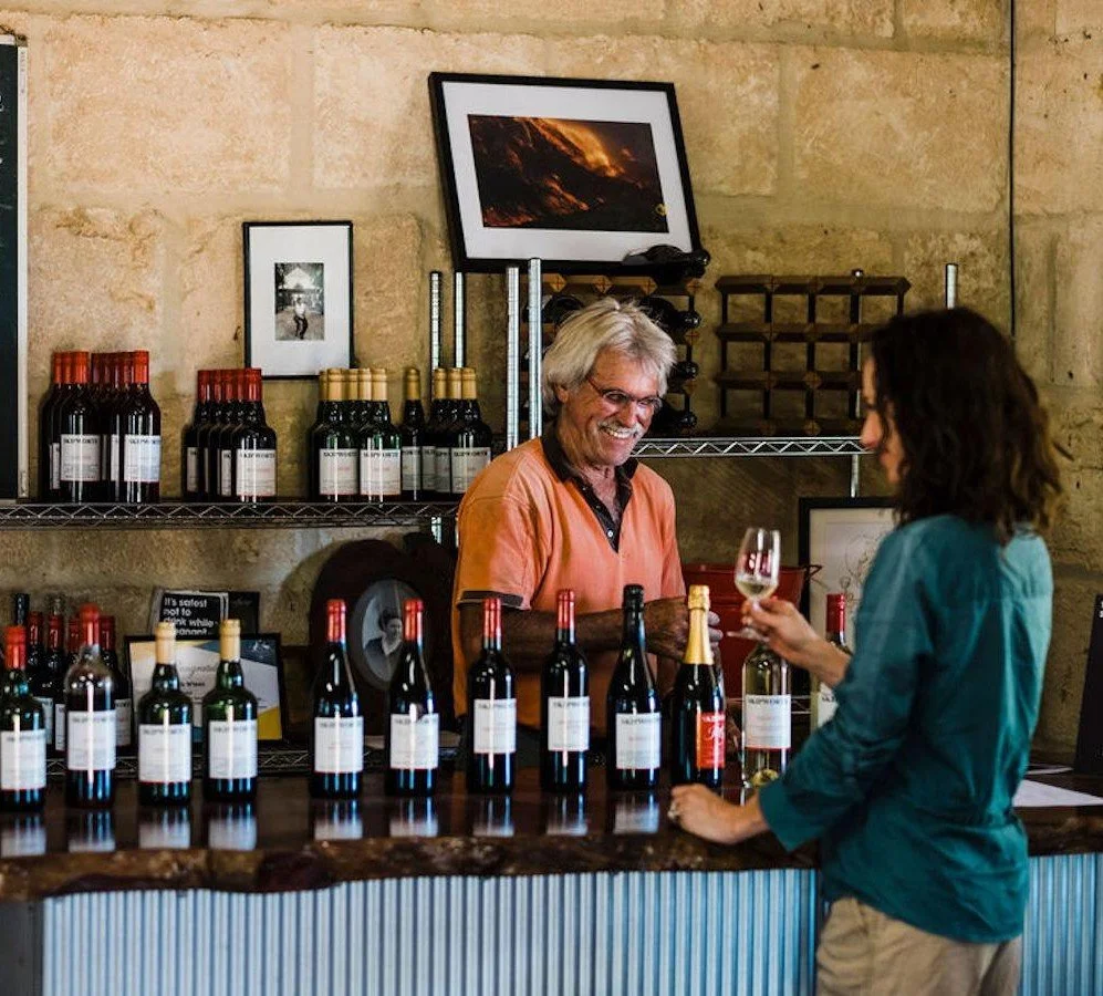 Kim Skipworth winemaker behind his wine tasting counter at the rustic cellar door of The Skipworth Wine Company, smiling and serving a lady in a teal shirt
