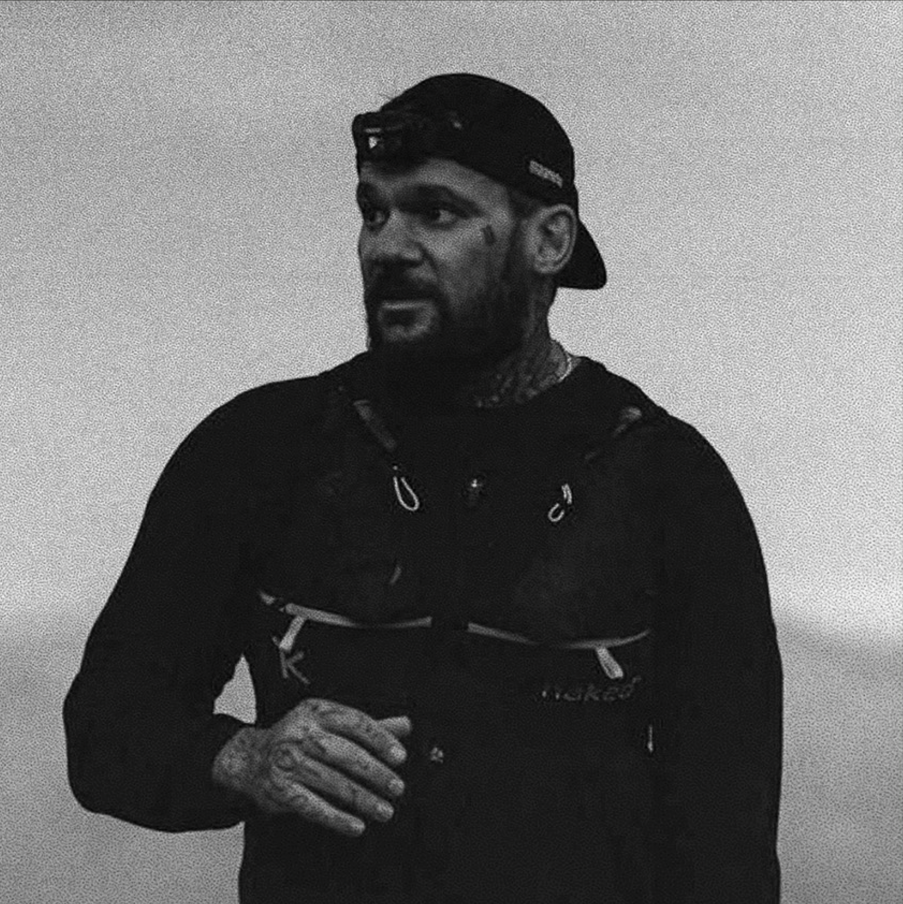 A man with tattoos and a beard wearing a backward baseball cap and athletic clothing, standing against a plain background.