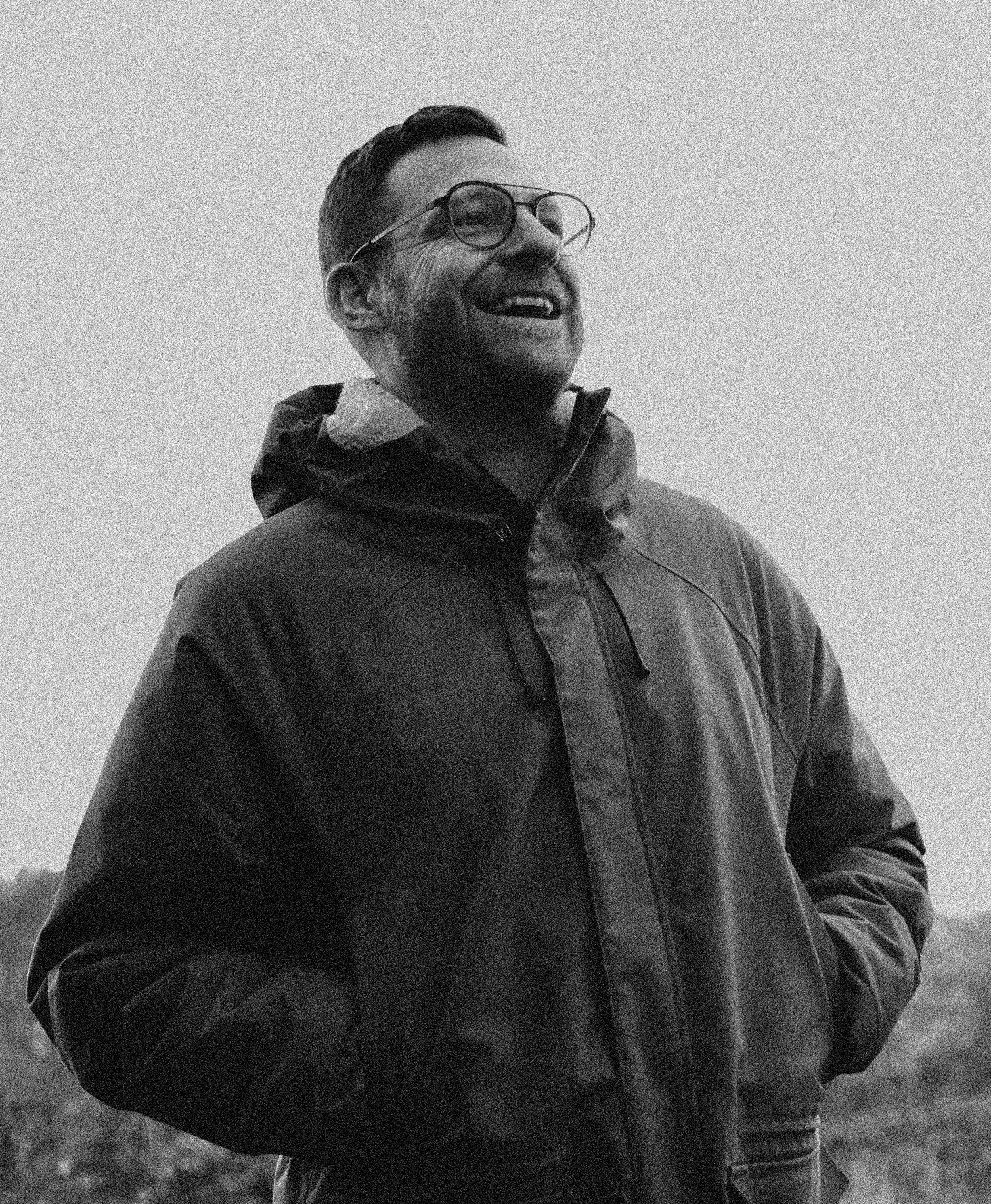 A laughing man with glasses and a beard, wearing a jacket, standing outdoors.