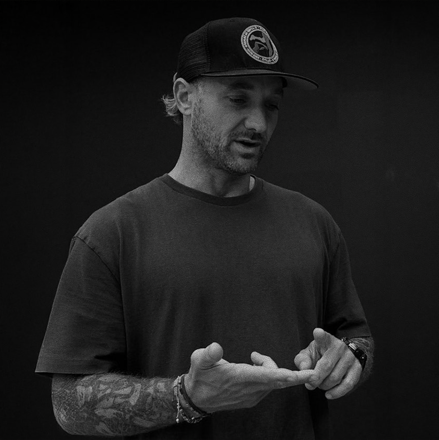 Black and white photo of a man wearing a baseball cap with a logo, a T-shirt, and bracelets, looking down and holding his hands together.