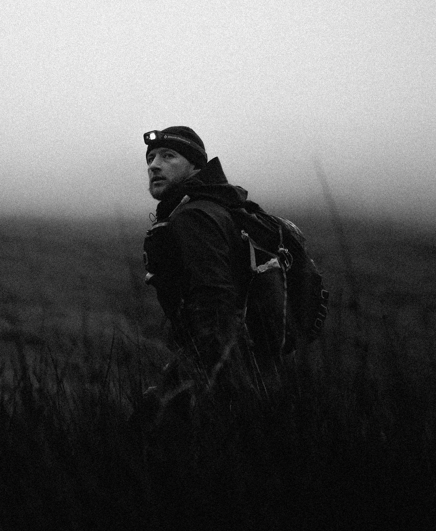 A man wearing a headlamp and outdoor gear with a backpack stands in a foggy, dark outdoor environment.
