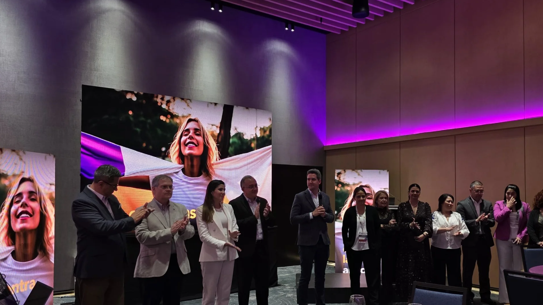 Varias personas en una conferencia o evento, de fondo pantallas con la cara de una mujer sonriendo con un fondo de árboles, iluminación púrpura y ambiente formal.