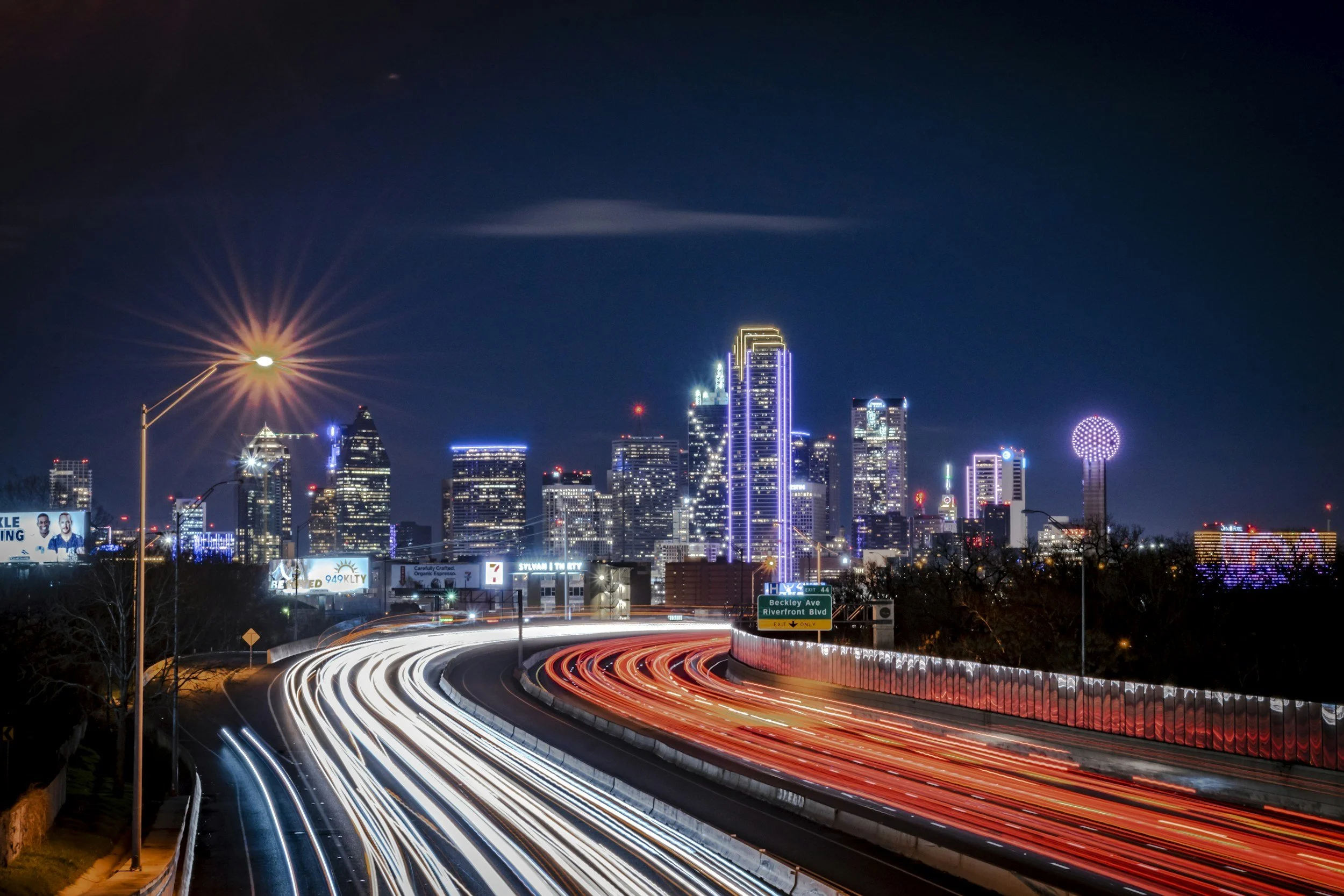Nighttime city skyline with tall illuminated buildings and light trails from moving vehicles on a curved highway in the foreground.
