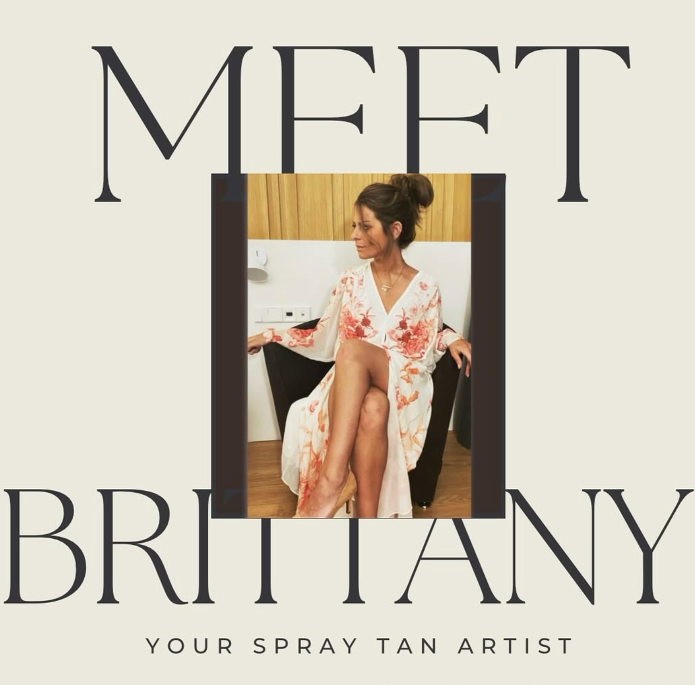 A woman with her hair in a bun wearing a floral robe, sitting on a black chair with her legs crossed, in a room with wooden walls, a white wall with electrical outlets and a lamp in the background.