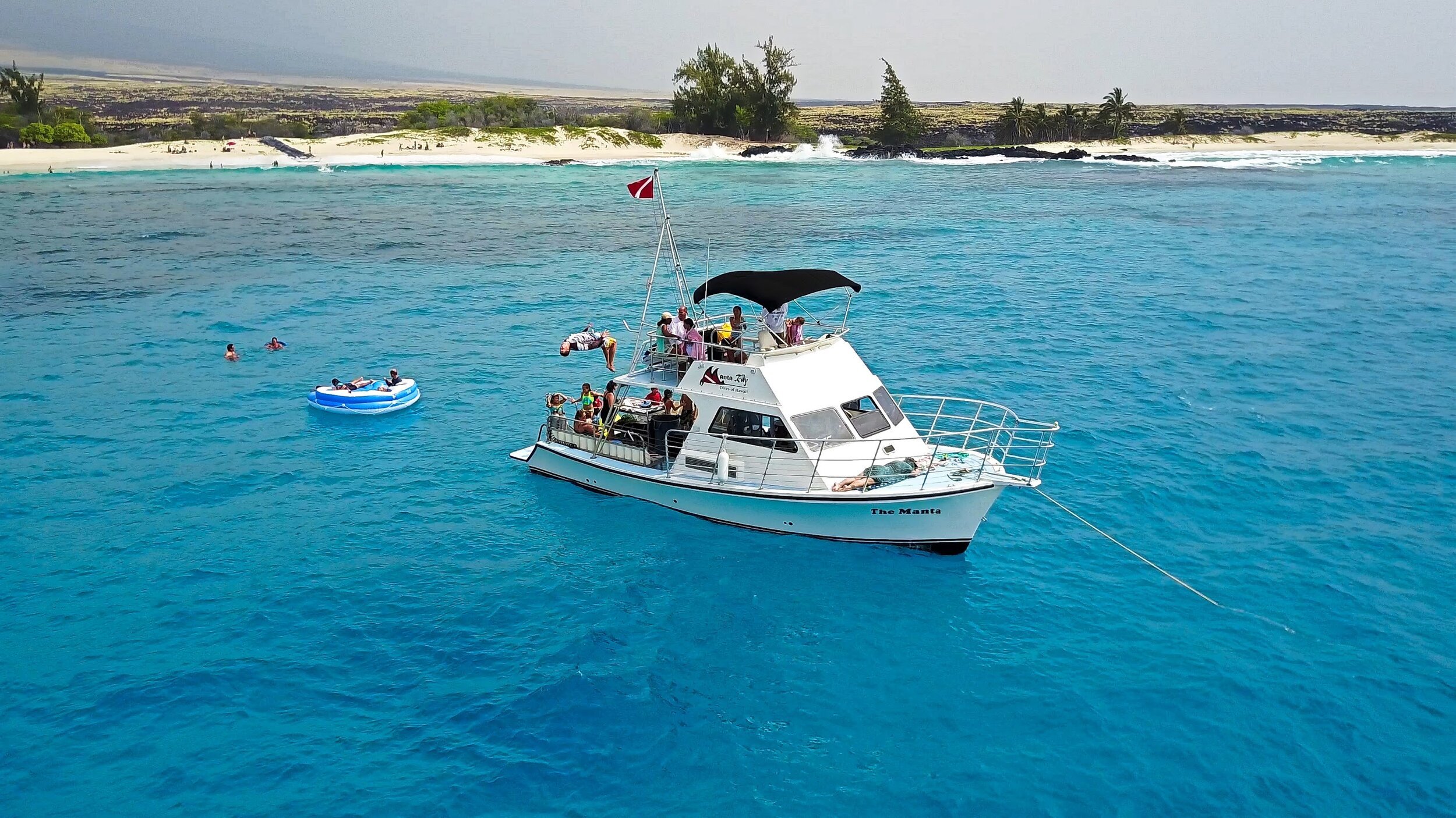 A white boat named The Manta anchored in clear blue water near a sandy island with sparse trees.