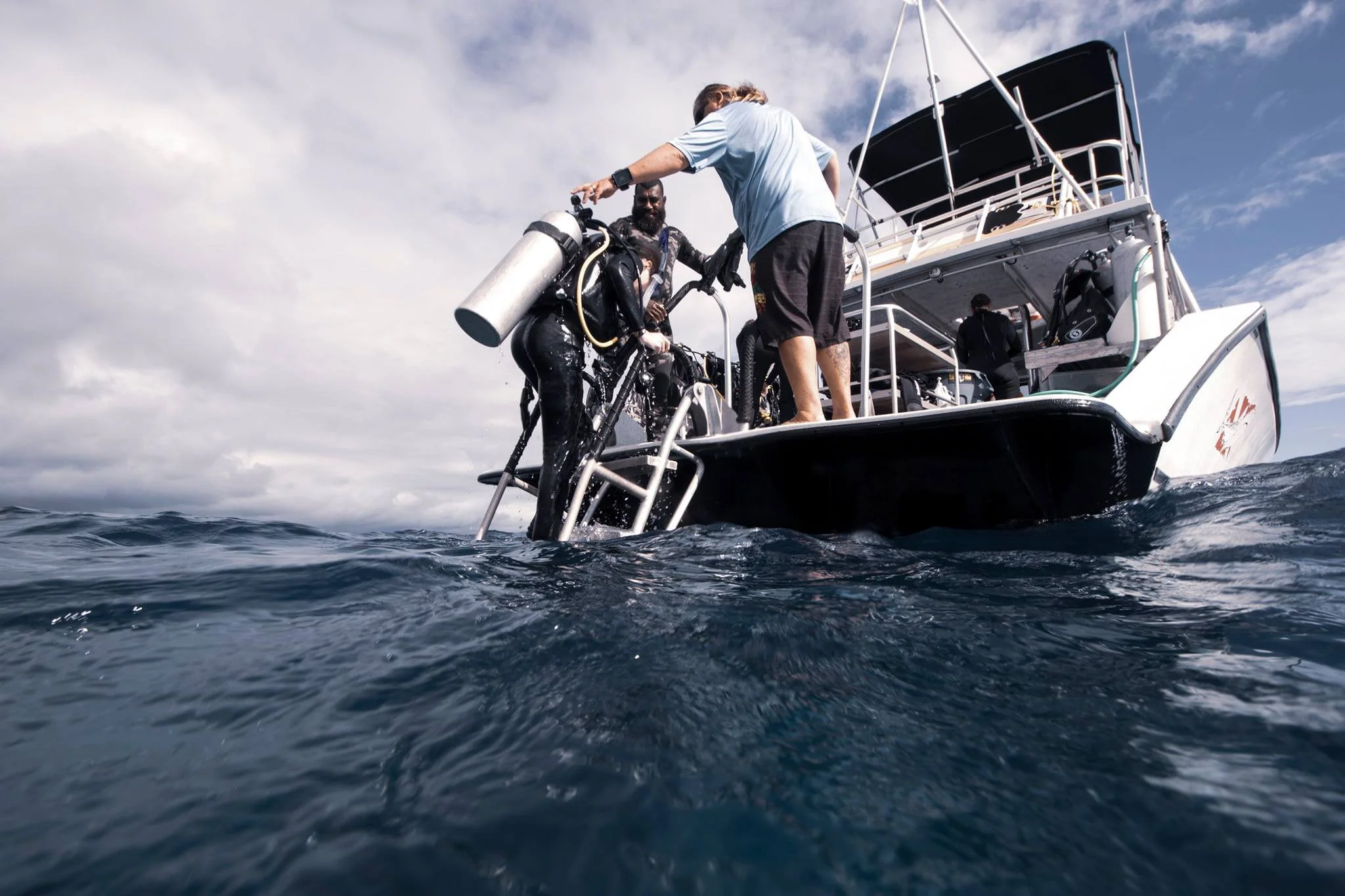 Three men on a boat preparing for scuba diving, with one man holding a bicycle and another man in scuba gear, in the open ocean under a cloudy sky.