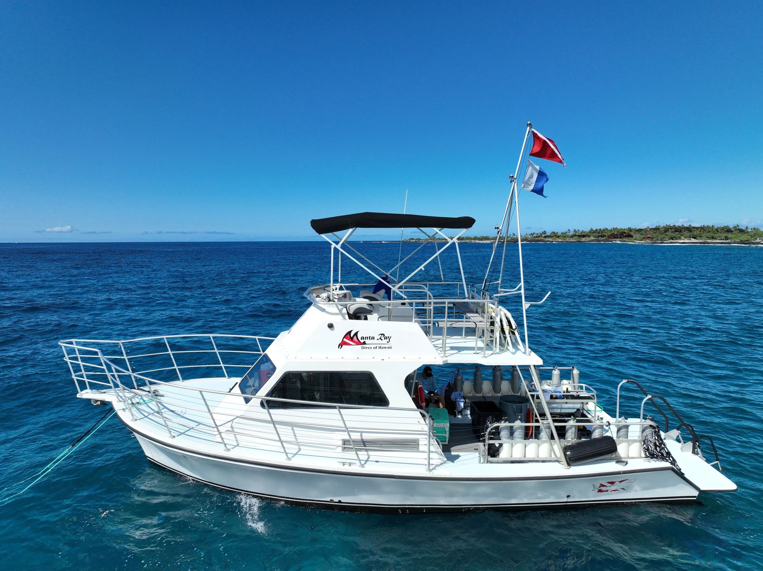 A white boat with a canopy, flying red and blue flags, on calm blue ocean waters near a distant shoreline with trees under a clear sky.