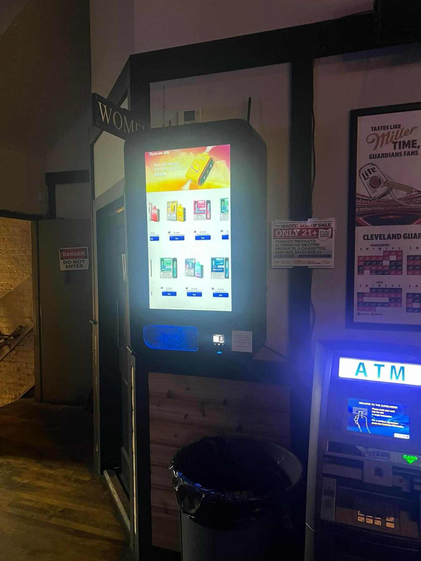 Vape vending machines installed next to an ATM in a nightlife venue, displaying age-restricted vape products and pricing in a dimly lit setting.