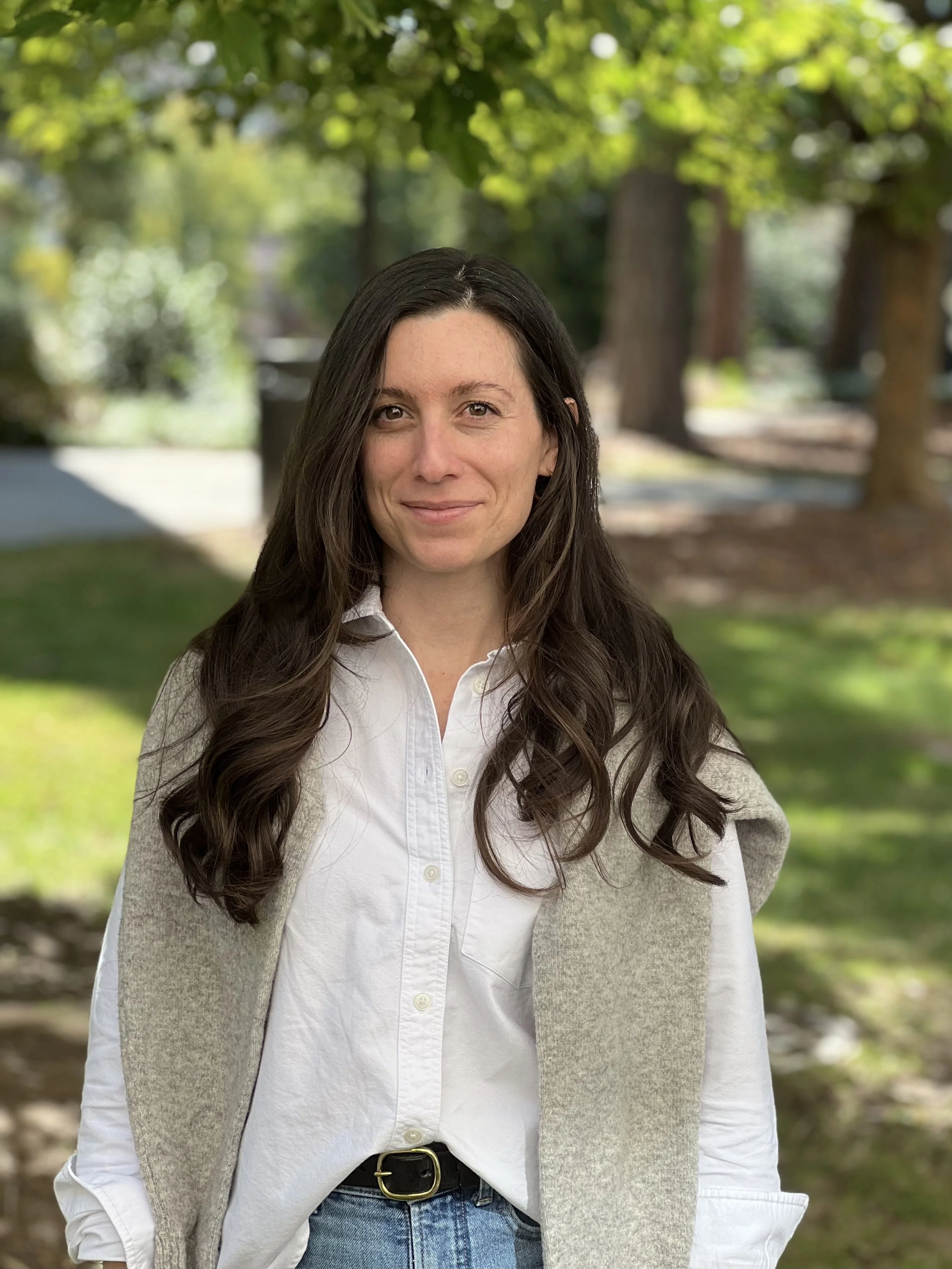 A woman with long dark hair smiling outdoors in a park with trees and grass.