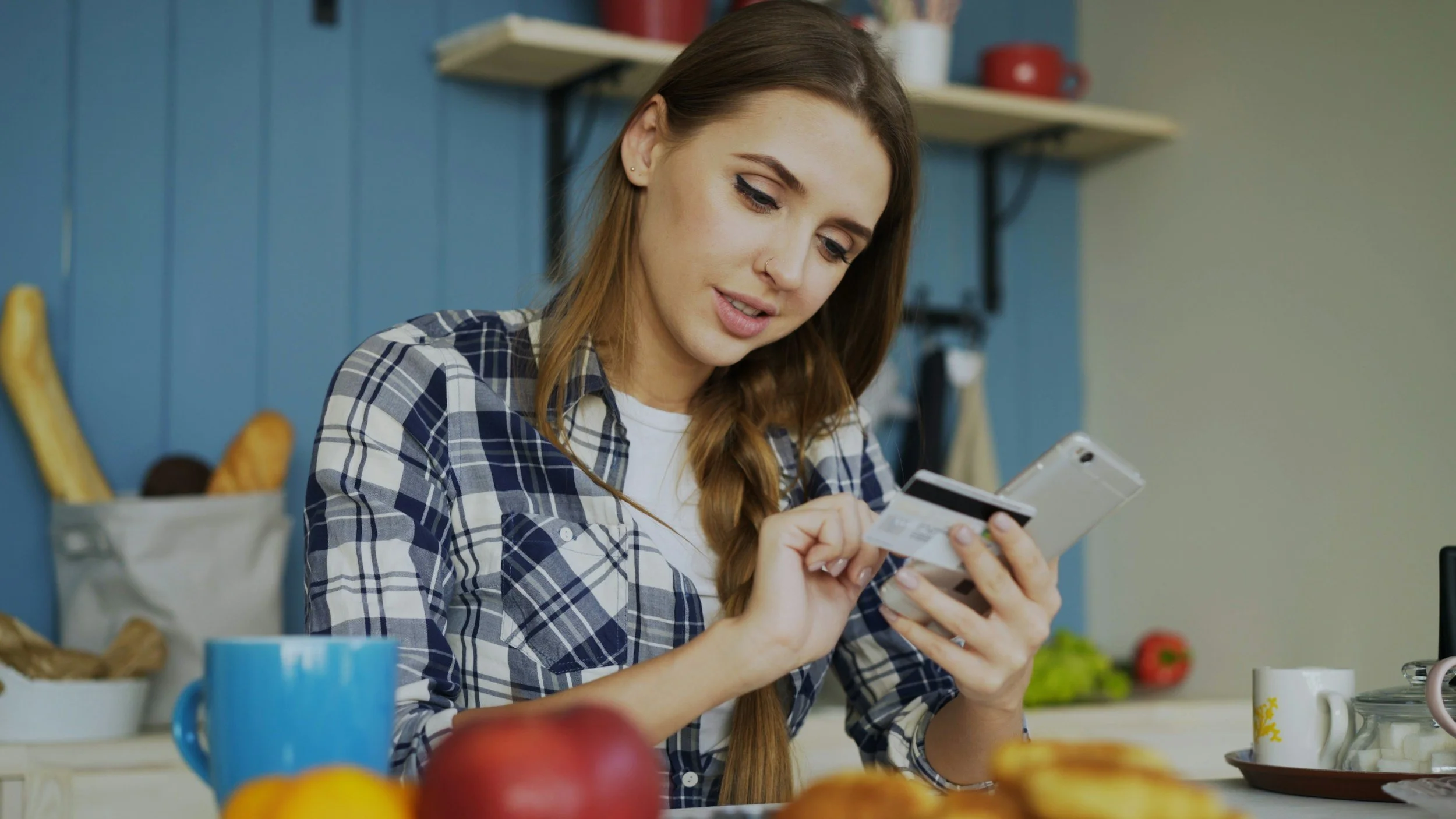 A young woman with long brown hair, wearing a plaid shirt, is looking at her phone and holding a credit card in a kitchen with a blue wall, shelves, and various food items.