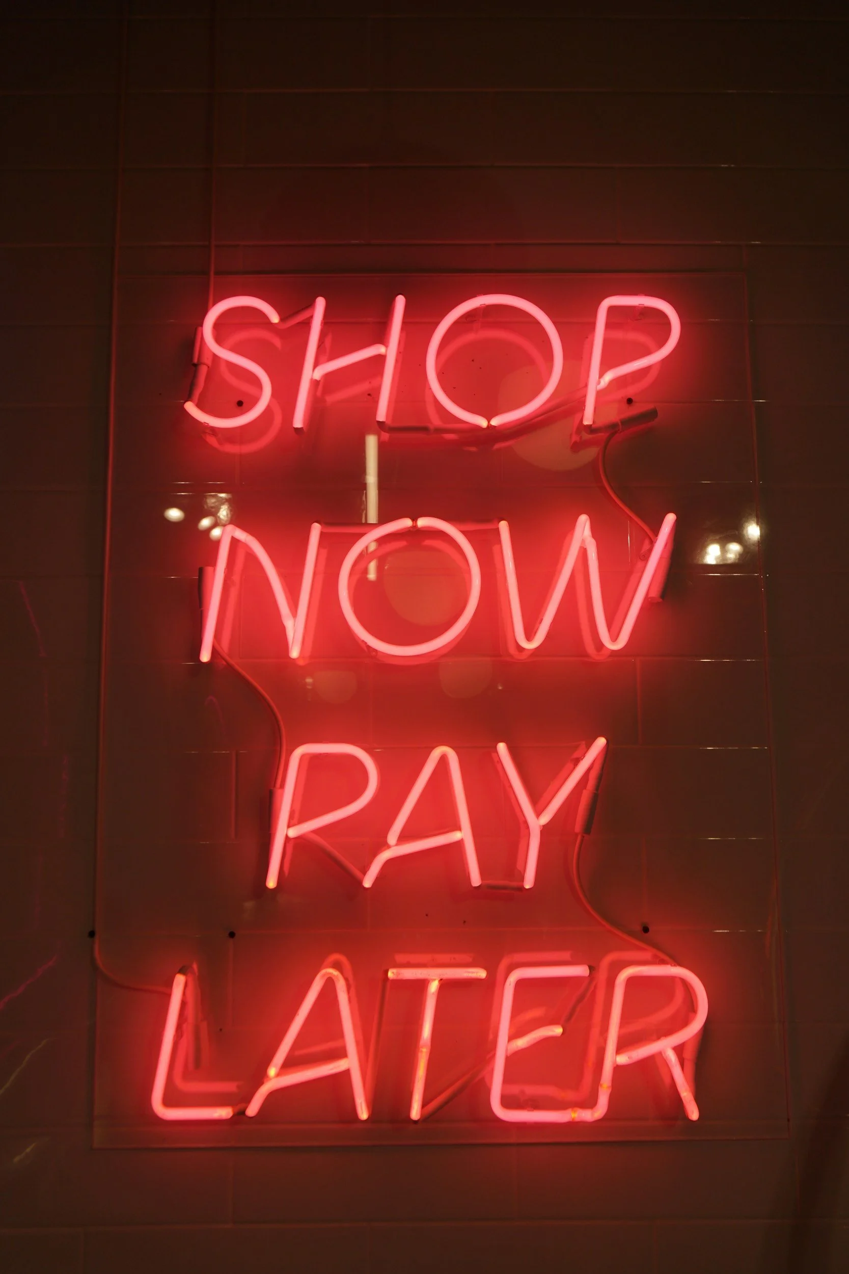 Pink neon sign with the message "SHOP NOW PAY LATER" on a dark wall.