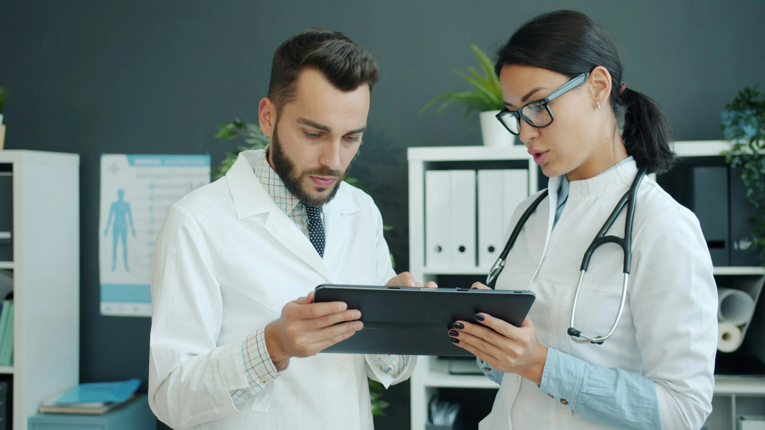 Two medical professionals, a man and a woman, in white coats looking at a tablet in a clinic or hospital setting.