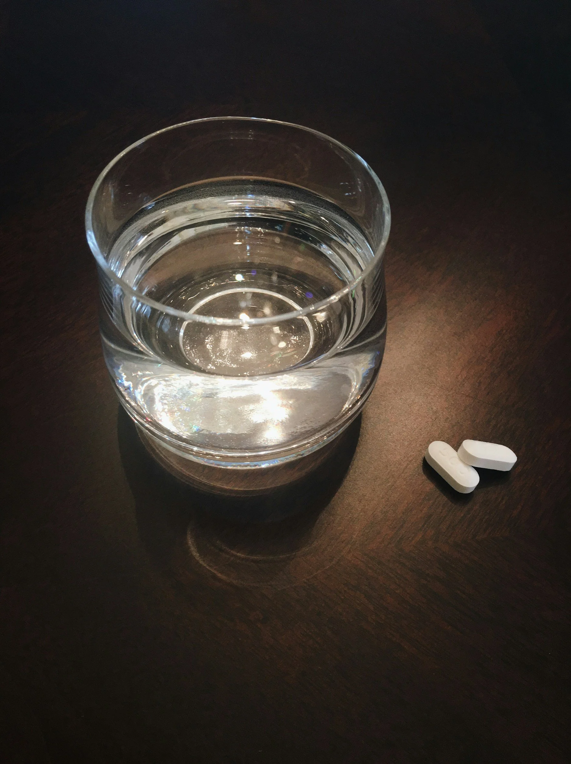 A clear glass of water on a dark wooden surface with two white pills nearby.