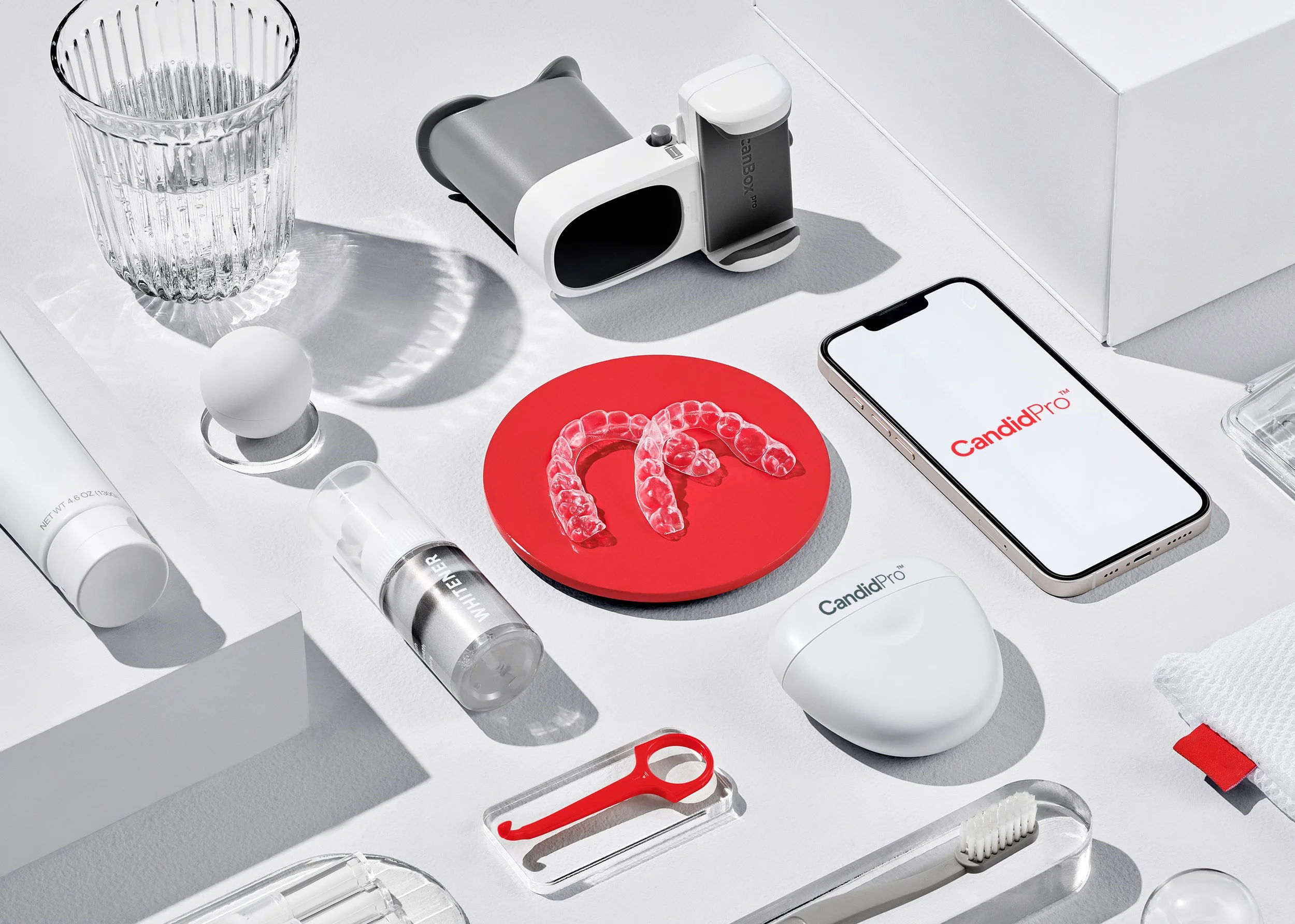 Dental impression trays with clear aligners on a red circular base on a white surface along with various dental tools, a smartphone displaying 'CandidPro', a glass of water, and other small dental-related items.