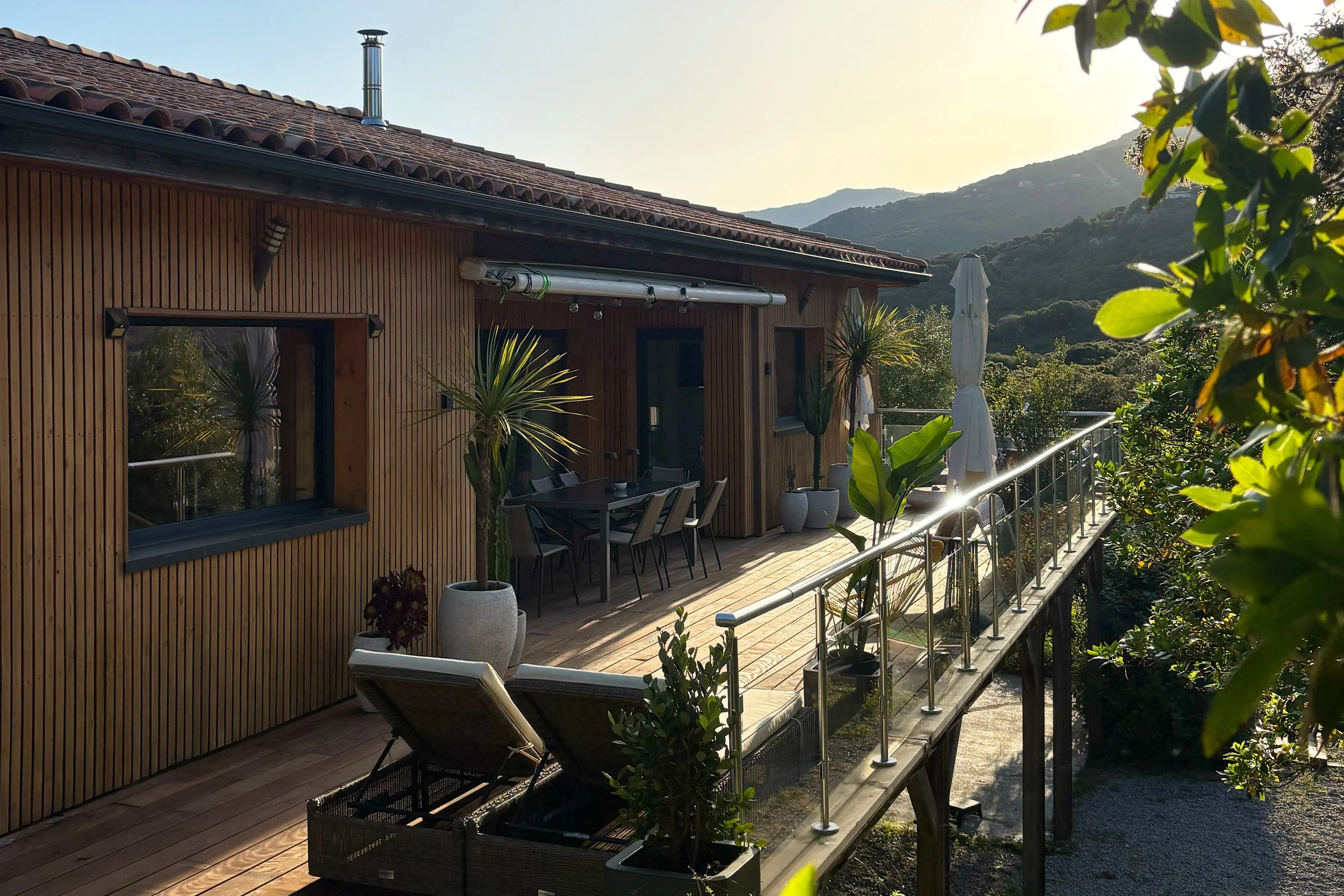 Vue d'une terrasse en bois avec des plantes en pot, une table avec des chaises, une grande parasol blanc, entourée de végétation et de montagnes au fond, sous un ciel ensoleillé.