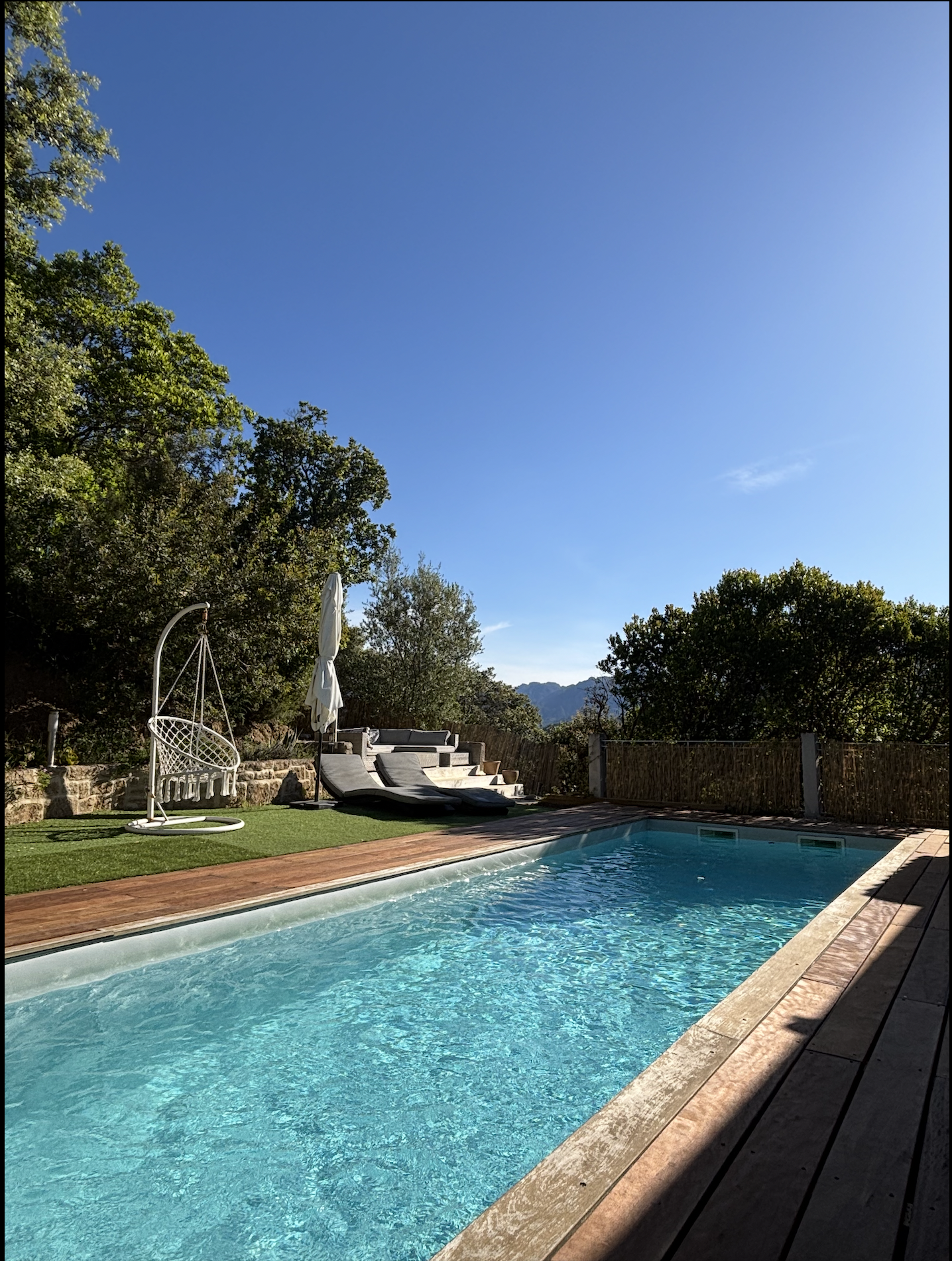 Une piscine dans un jardin entouré d'arbres, avec des chaises longues, un parasol fermé, un mobilier de détente et un ciel bleu clair.