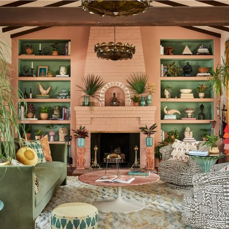 Colorful living room with a pink brick fireplace, green bookshelves, and bohemian decor. There are plants, statues, and books on the shelves, and patterned chairs and a green sofa around a pink round coffee table.