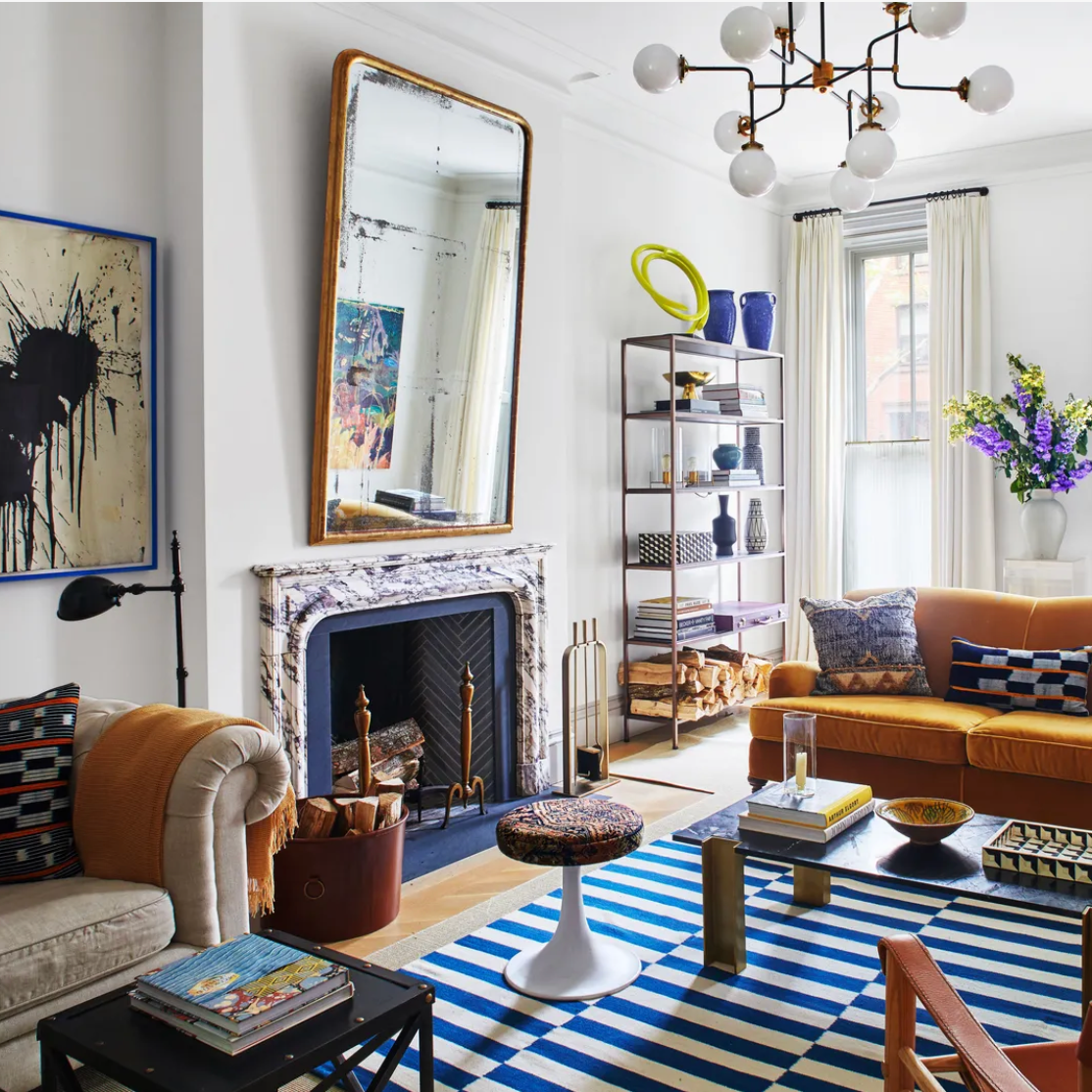 A well-decorated living room with a mustard yellow couch, a marble fireplace, a large standing mirror above the fireplace, a modern bookshelf with vases and books, a striped blue and white rug, a small multicolored stool, a dark coffee table with magazines and a chessboard, and windows with cream curtains, decorated with purple and yellow flowers in a white vase.