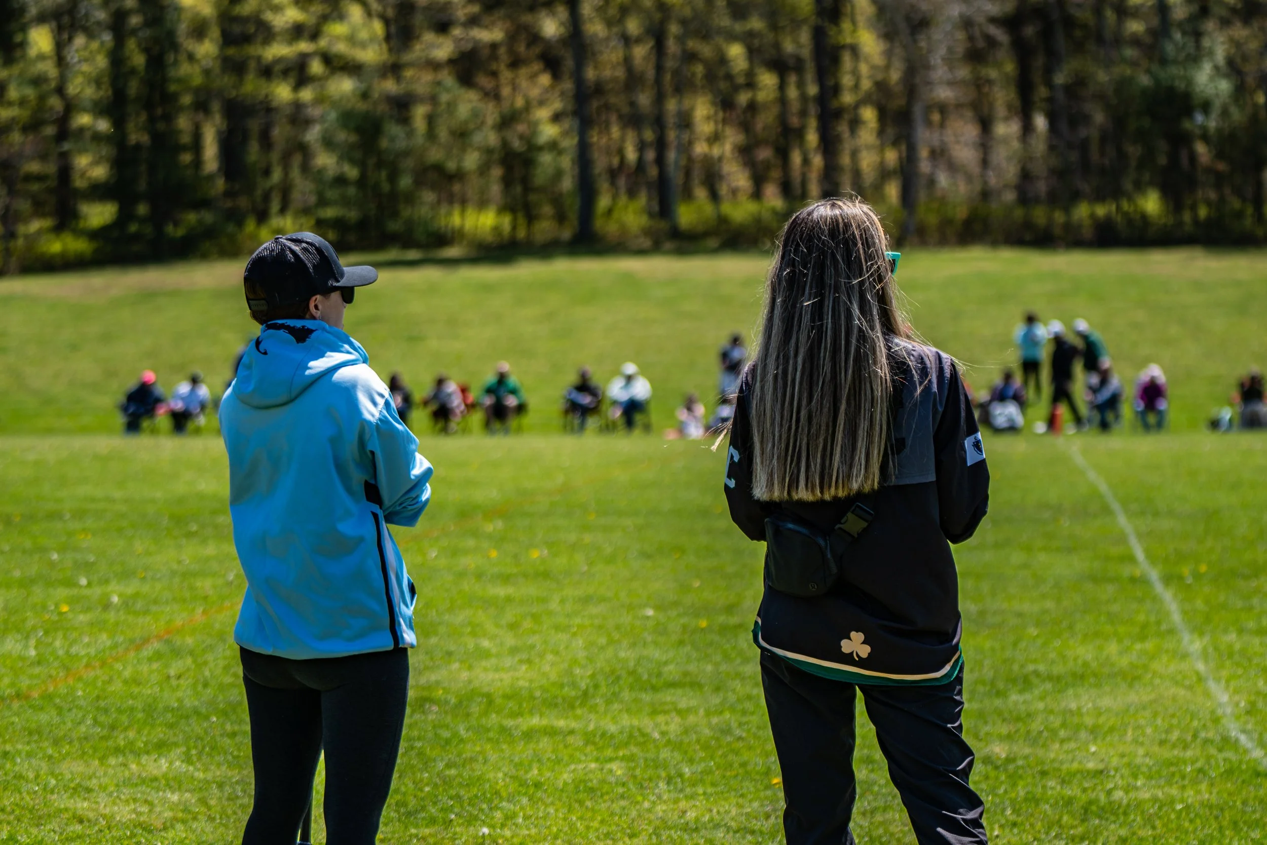 People sitting and standing on the grass field of a park with trees in the background