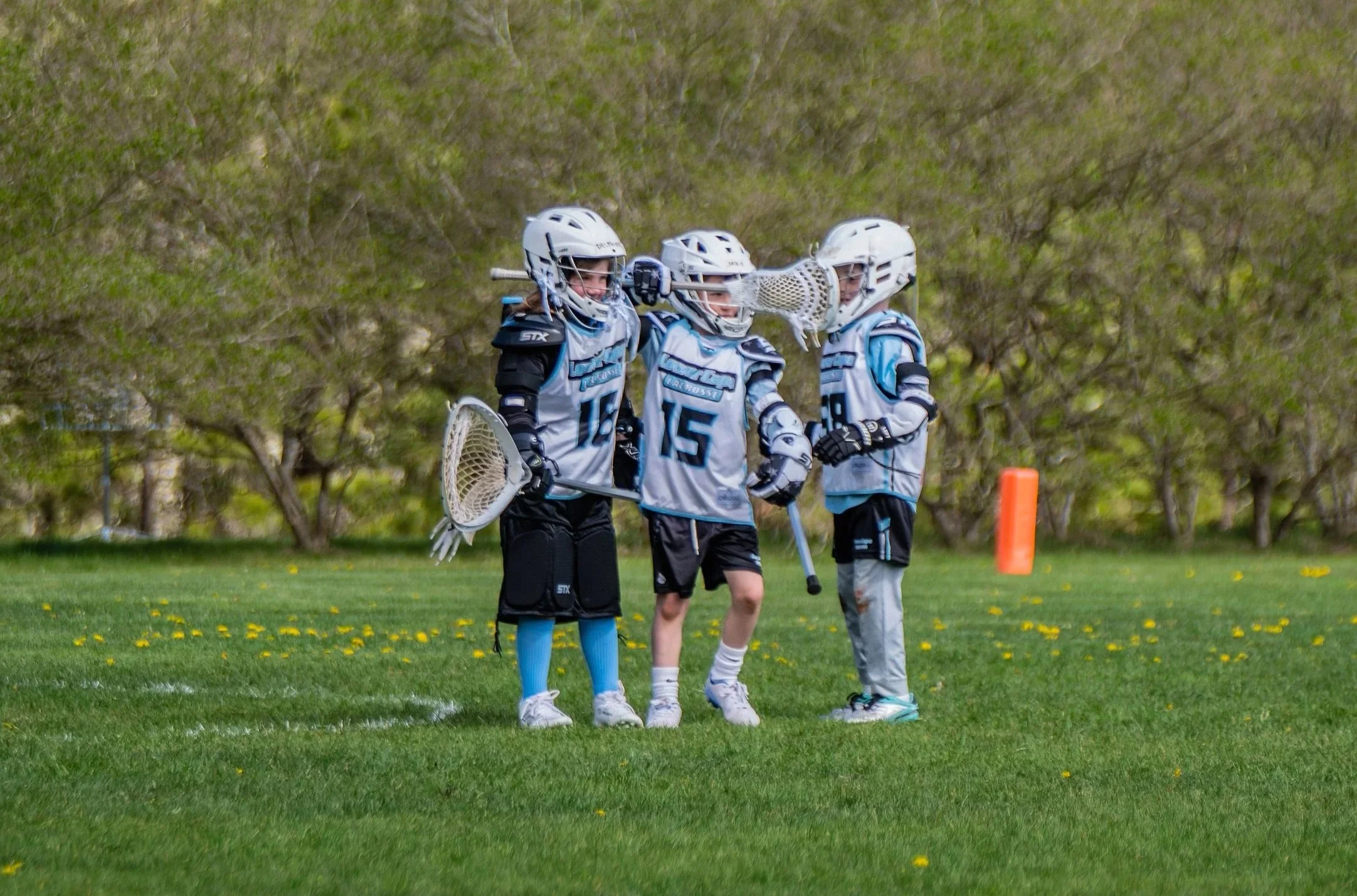 Three young lacrosse players wearing uniforms and helmets standing on a grassy field, engaging in a team discussion during a game.
