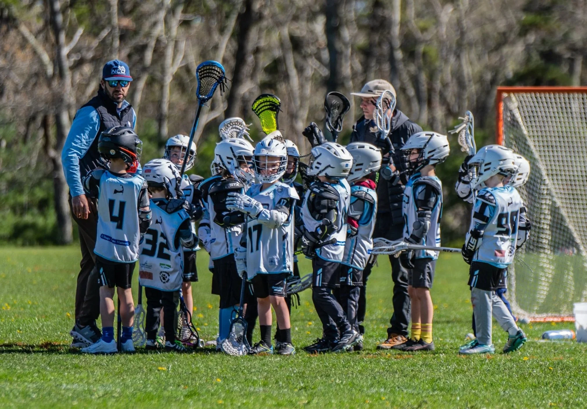 Lacrosse coach instructing young players on a grassy field with trees in the background. The children wear helmets and jerseys, holding lacrosse sticks.