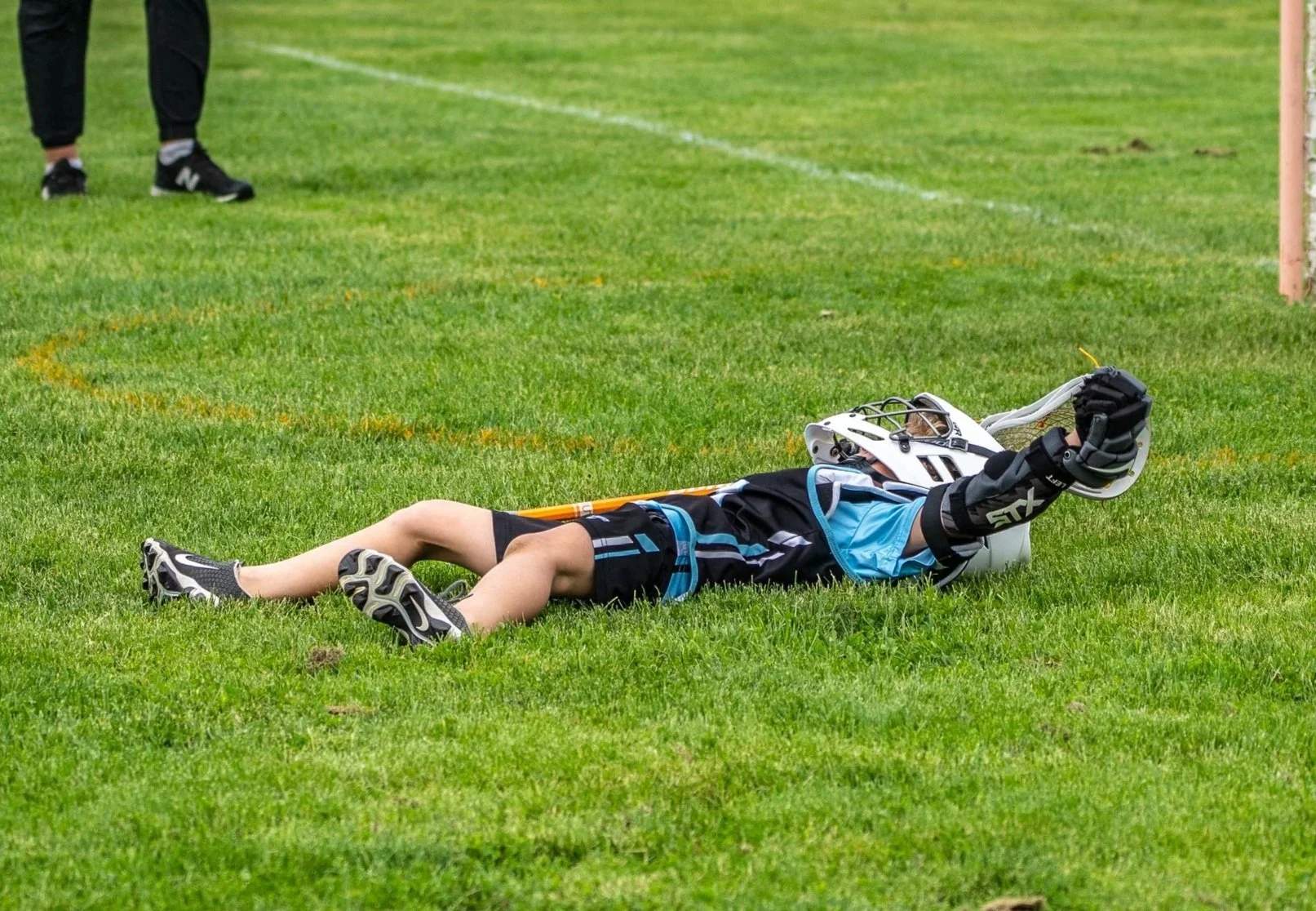 A child lying on the grass wearing a hockey helmet and gloves, holding a hockey stick.