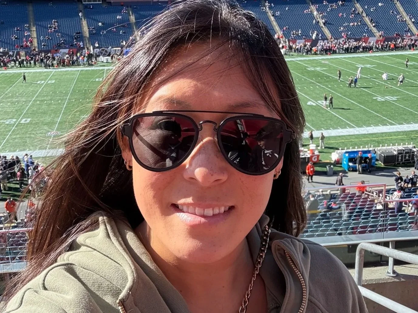 A woman wearing sunglasses and a hoodie taking a selfie in a stadium with a football field in the background.