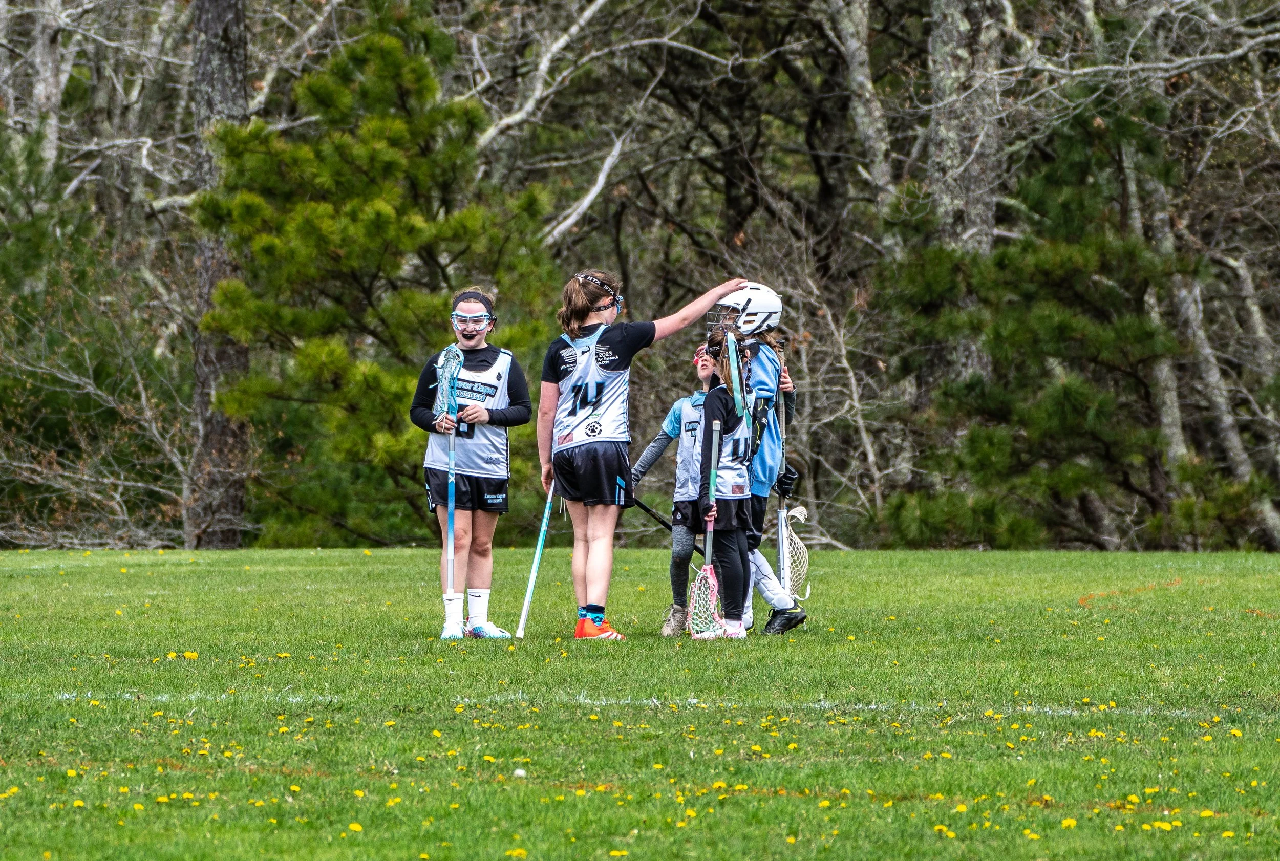 A group of young girls playing lacrosse on a grassy field, with some handing their helmets and sticks to each other, and trees in the background.