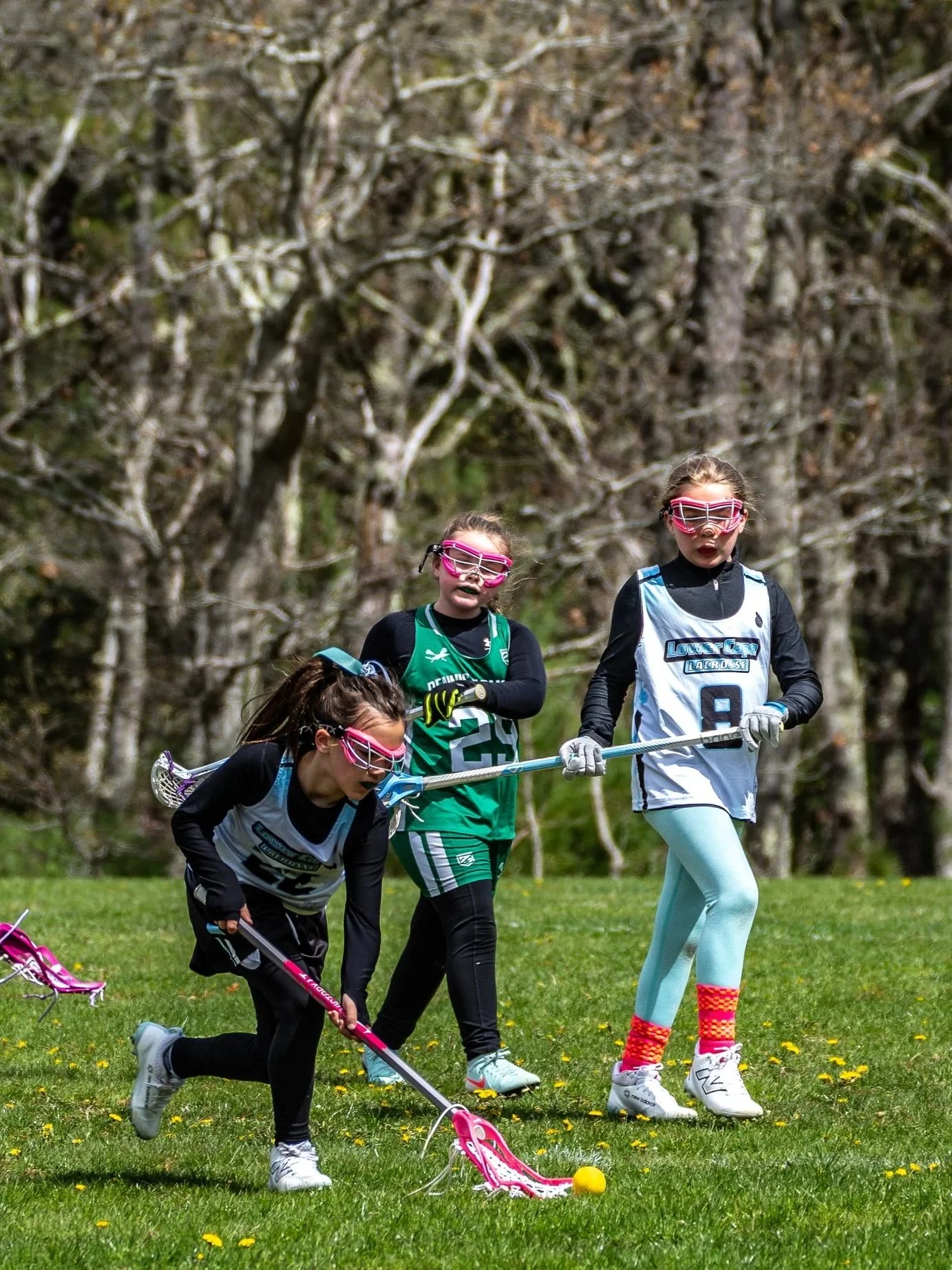 Three young girls playing lacrosse outdoors on a grassy field with trees in the background. They are wearing sports gear and goggles, with one girl crouched down handling a lacrosse stick and the other two standing nearby with their sticks.