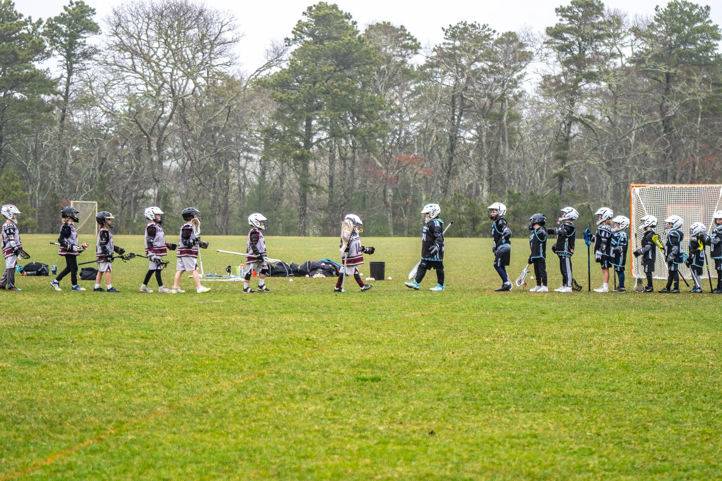 Kids playing lacrosse on a grass field, divided into two teams, with many trees in the background.