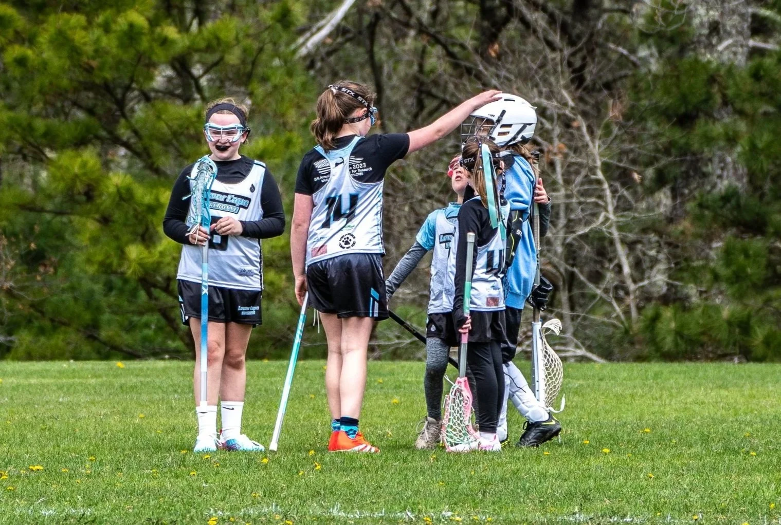 A group of young girls in sports uniforms with lacrosse sticks, gathered outdoors on a grassy field, with one girl adjusting a helmet on another.