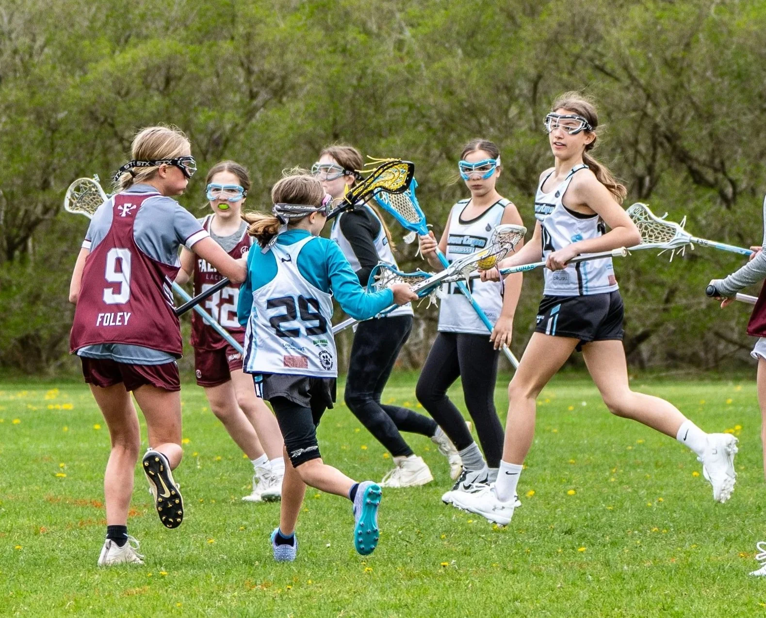 Young girls playing lacrosse on a grassy field in a group huddle, wearing sports uniforms and protective goggles, holding lacrosse sticks.