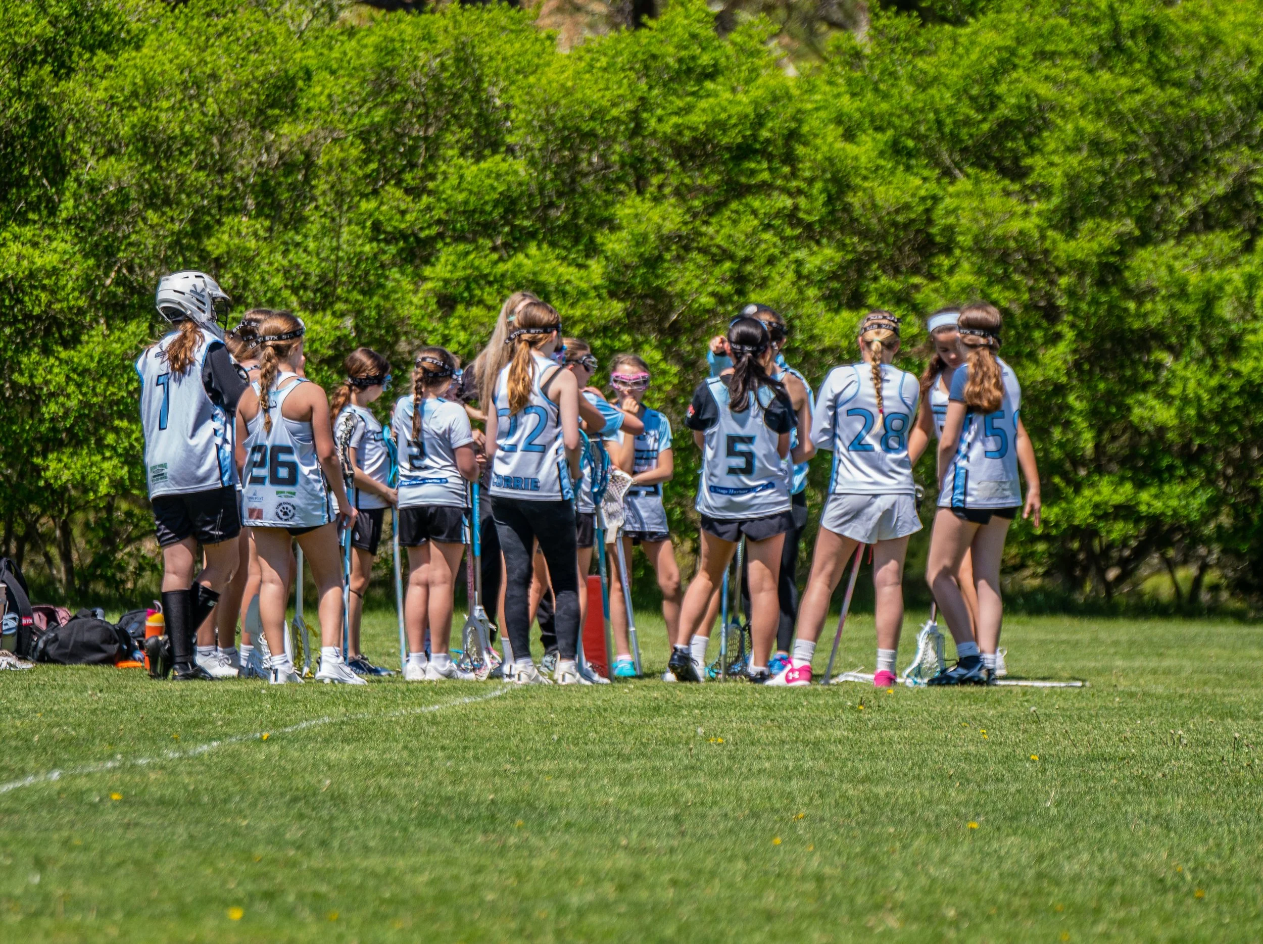 A group of young girls in sports uniforms and protective gear, standing on a grassy field with lush green bushes in the background, appears to be a lacrosse team huddle.