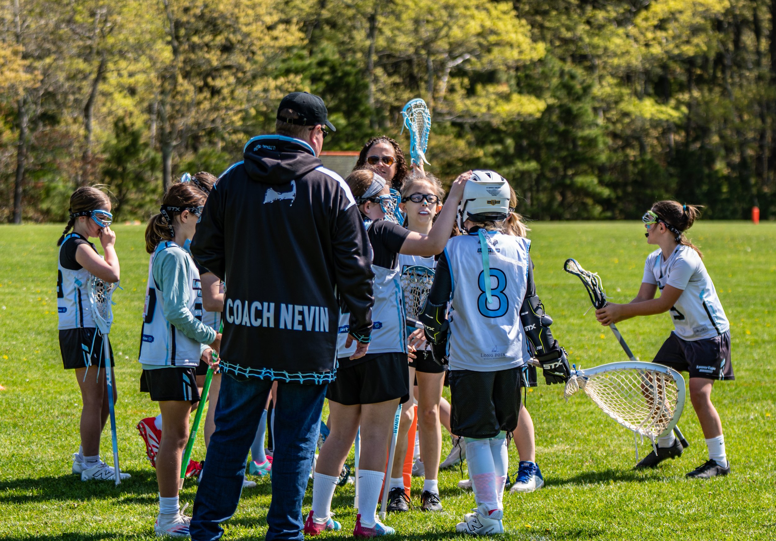 Girls' lacrosse team huddled on a grassy field with coach Nevin, who is wearing a black jacket with