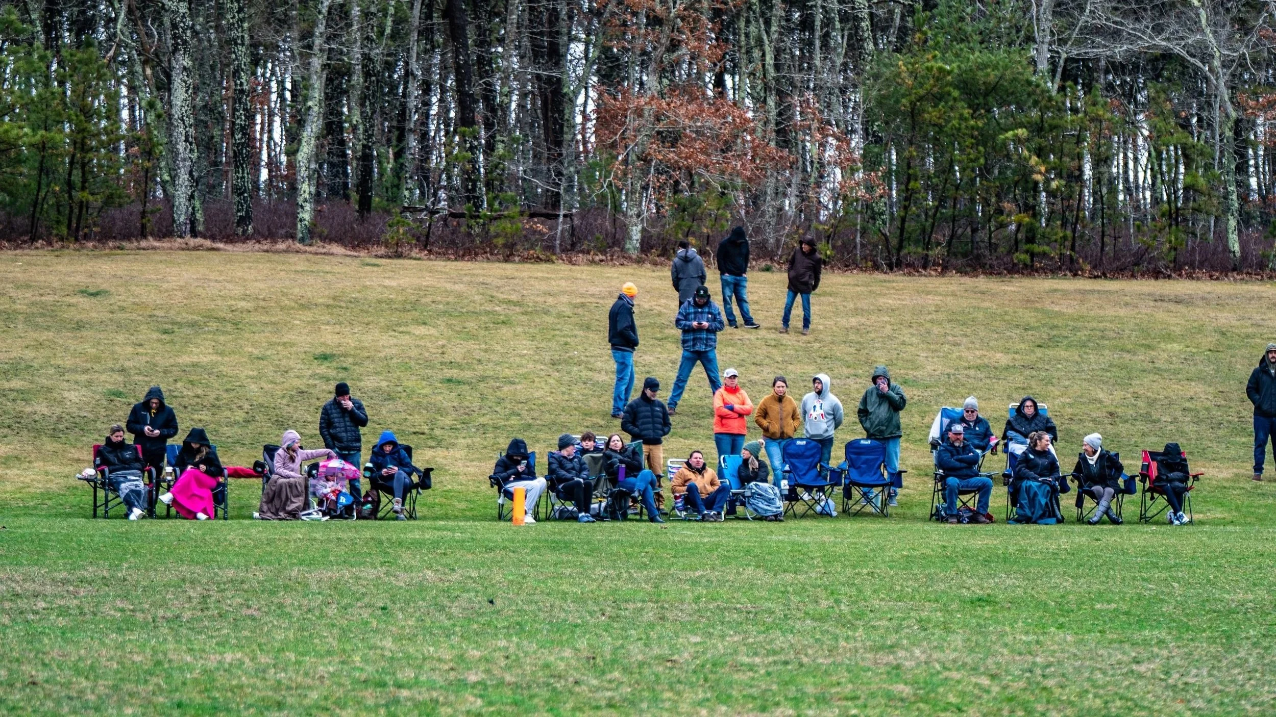 A group of people sitting and standing on a grassy field near a wooded area, some seated in camping chairs and others standing, dressed in warm clothing indicating cool weather.