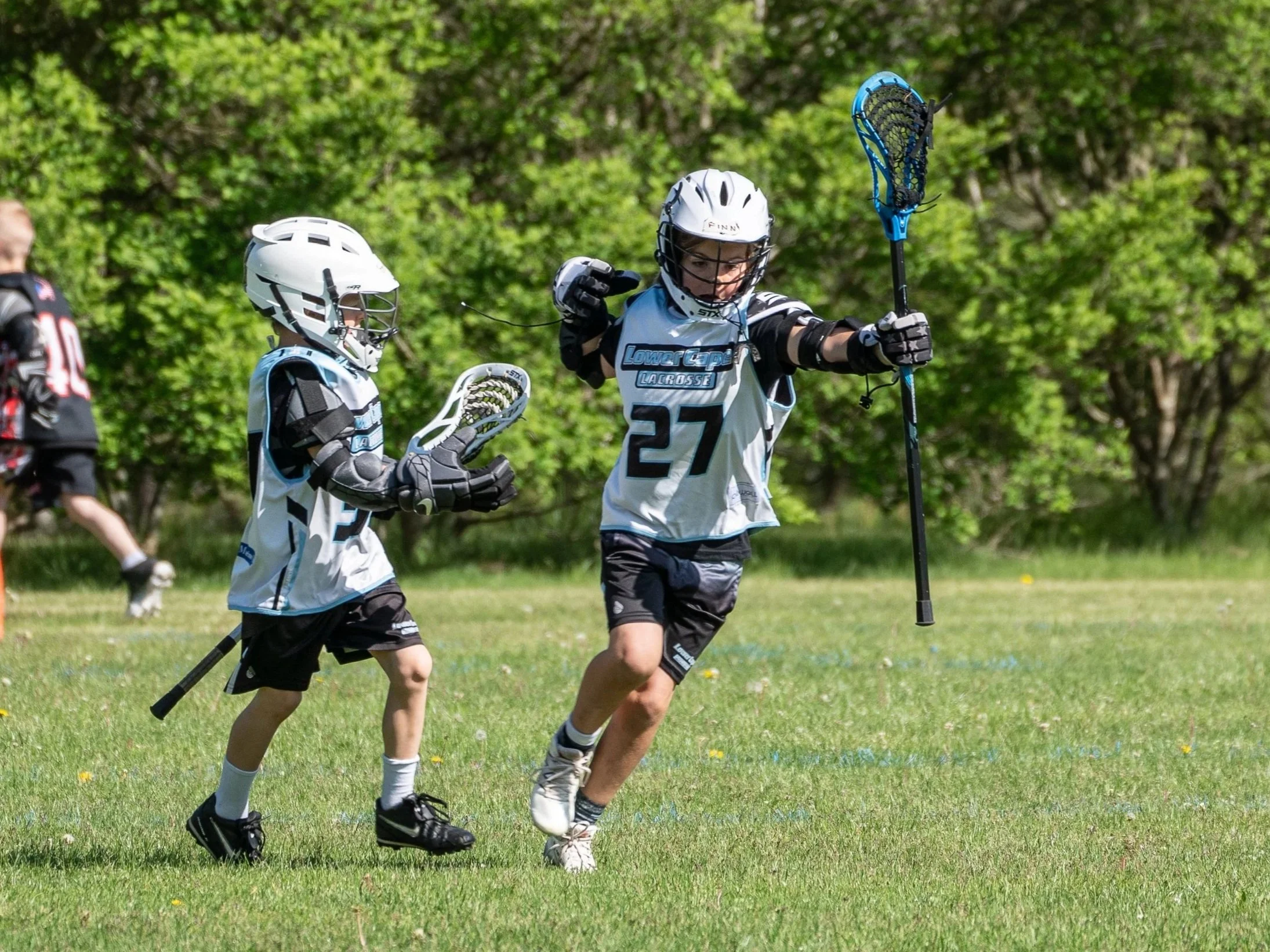 Two young lacrosse players in white and black uniforms with helmets, gloves, and pads playing on a grassy field with green trees in the background. One is running holding a lacrosse stick, the other is preparing to catch or pass the ball.